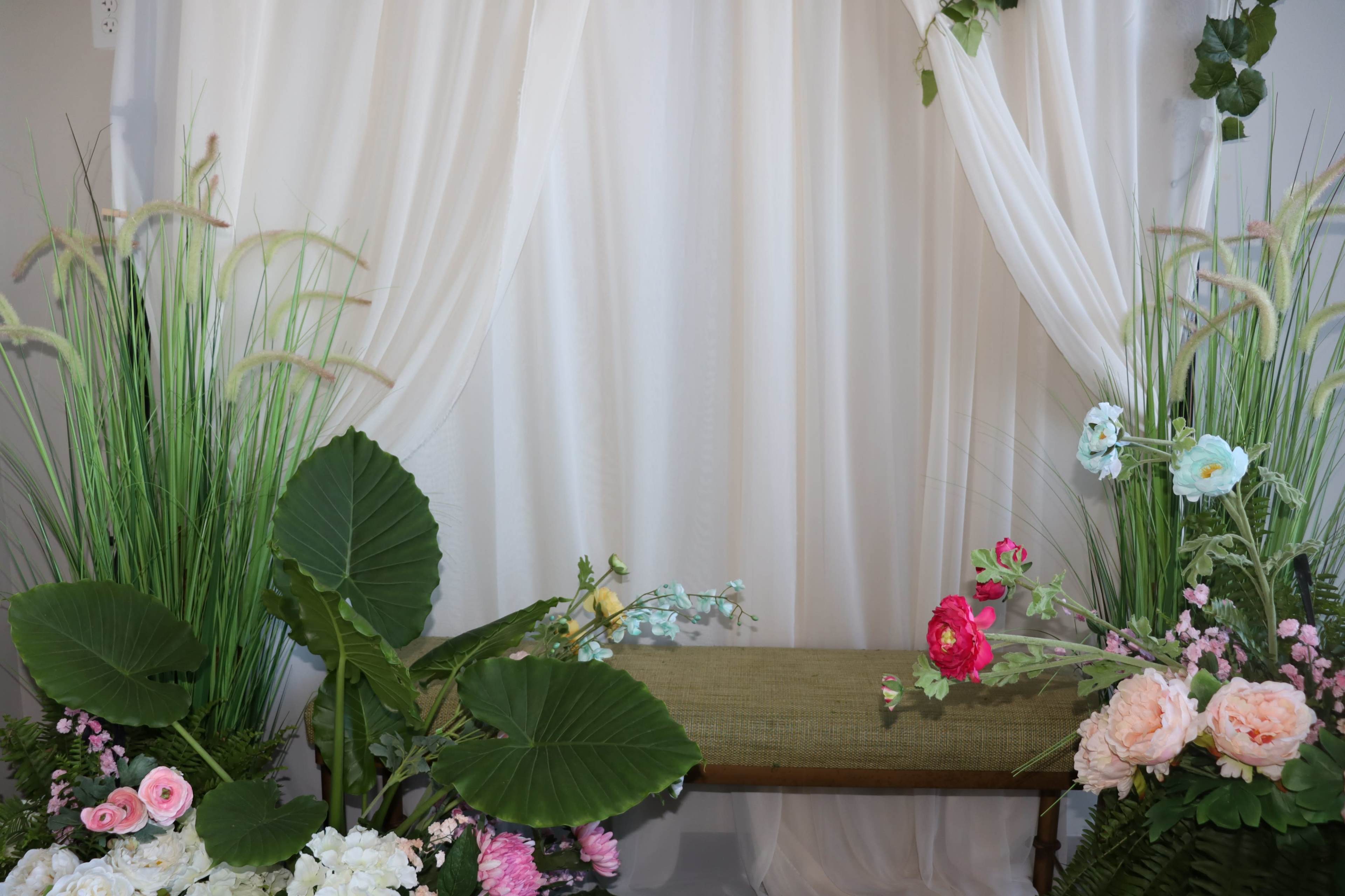A decorative setup with a floral arrangement on each side of a wooden bench and a white curtain backdrop.