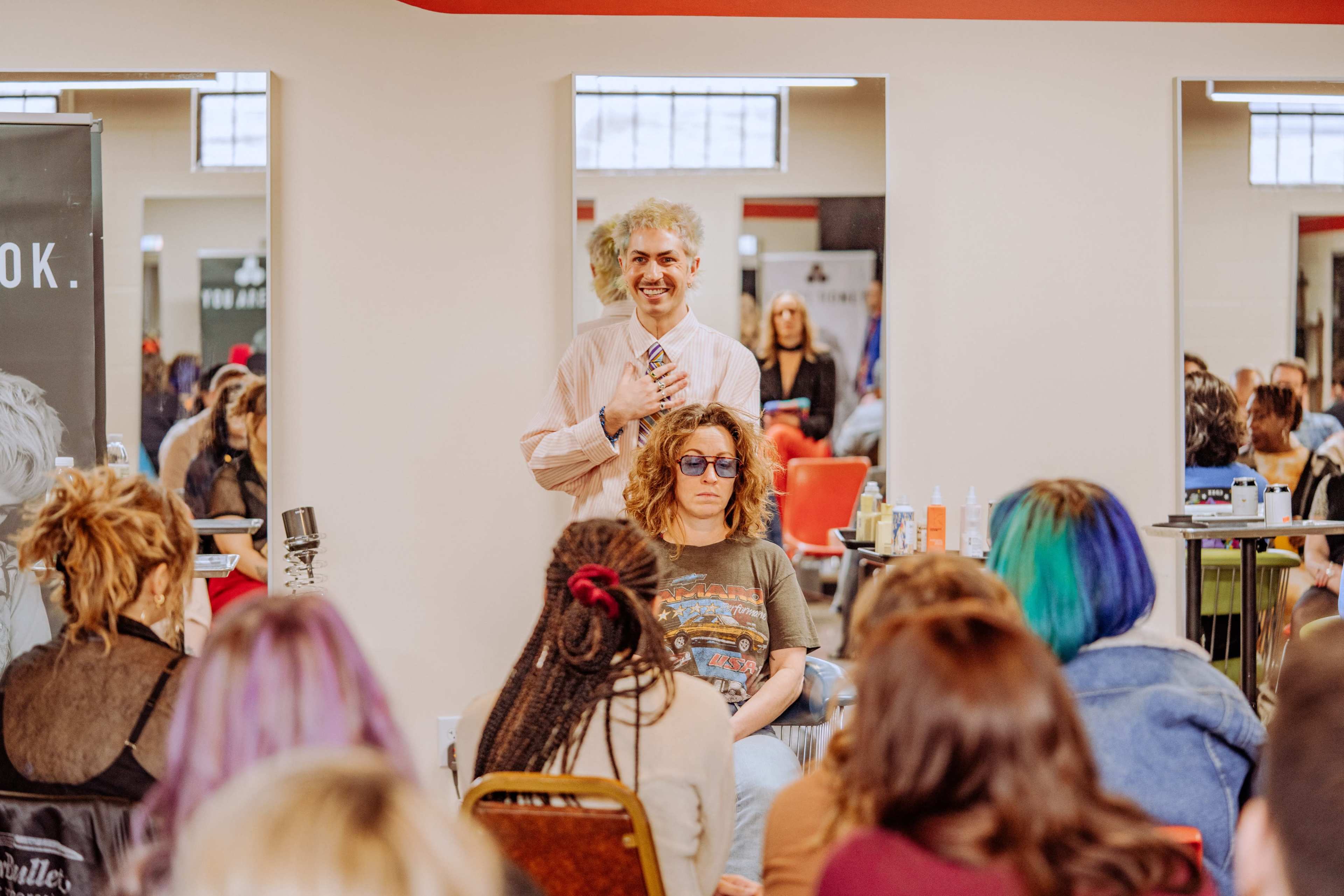 A man stands in front of a seated woman with curly hair and glasses while a group of people observes in a well-lit salon setting.