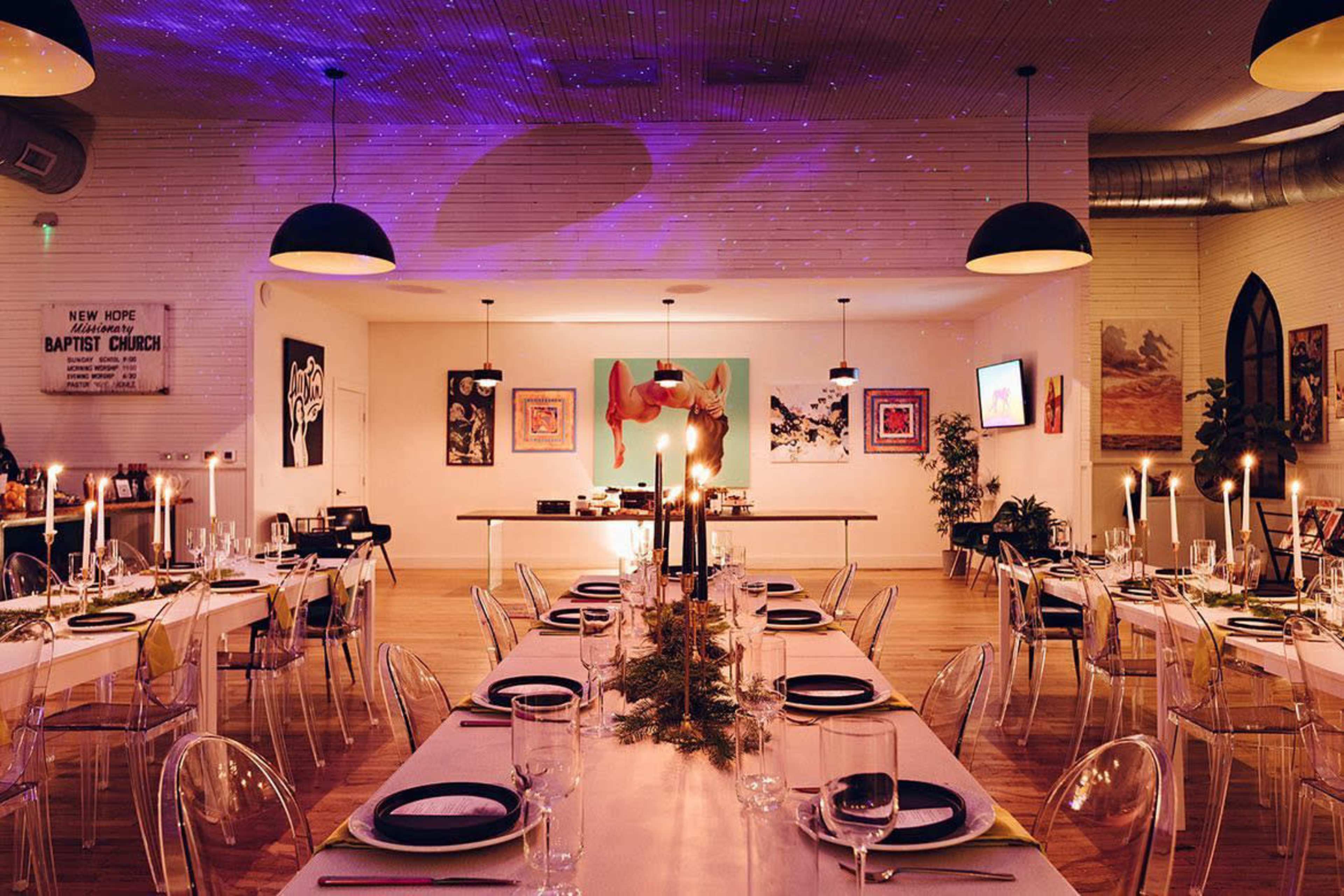 A spacious dining room features long tables set for a meal, adorned with candles and centerpieces, under softly colored lighting and decorative artwork.