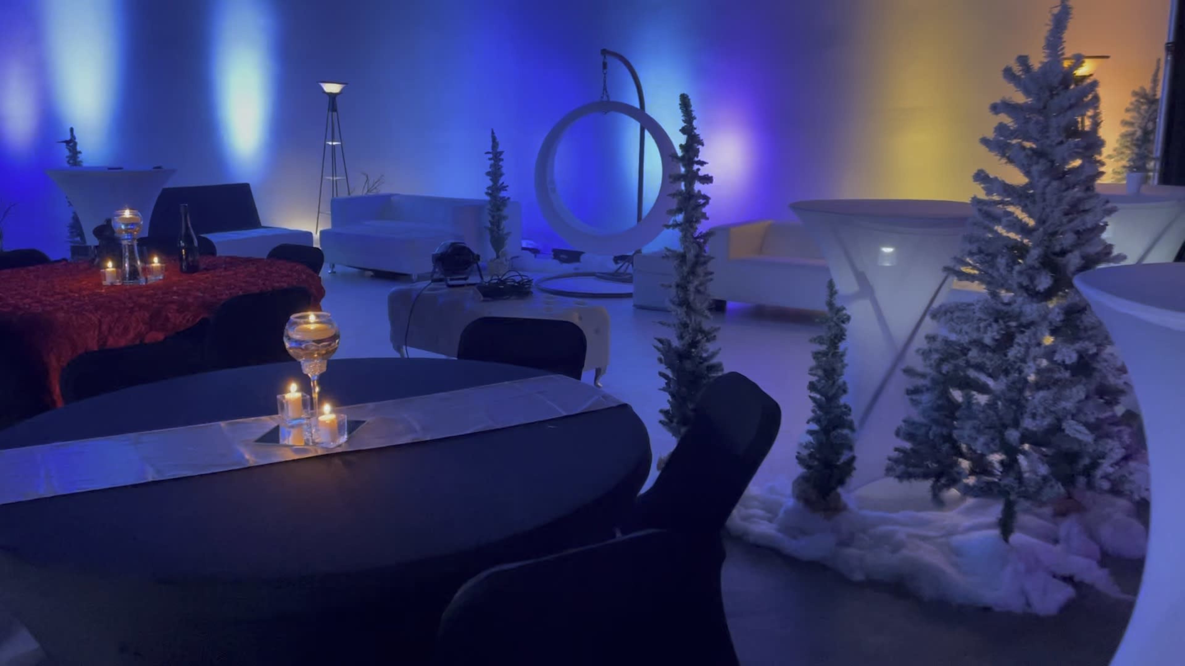 A decorated event space with tables, candles, and artificial snow-covered trees, illuminated by colorful lights.