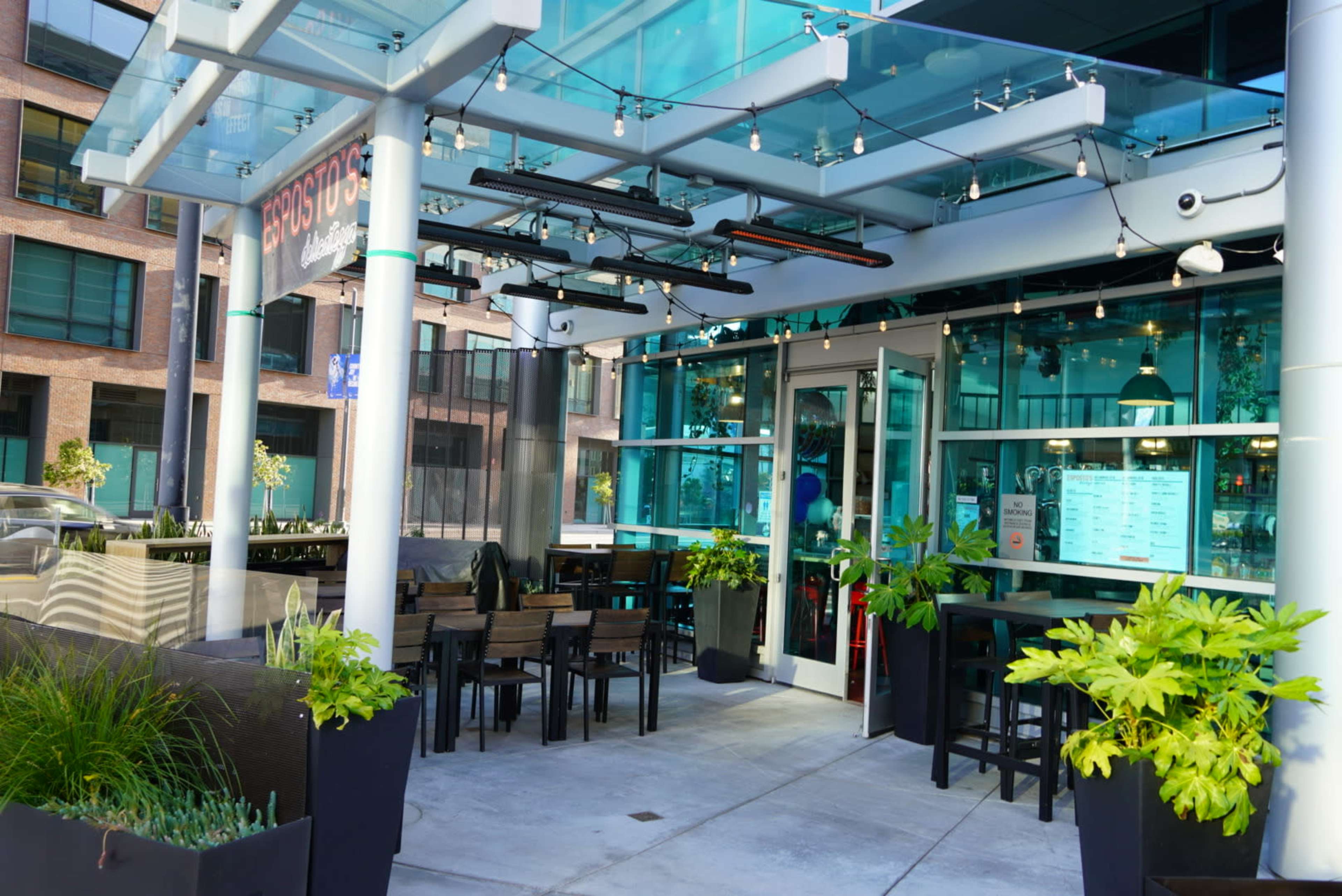 An outdoor seating area of a modern restaurant featuring tables and chairs under a glass canopy with plants and lighting fixtures.