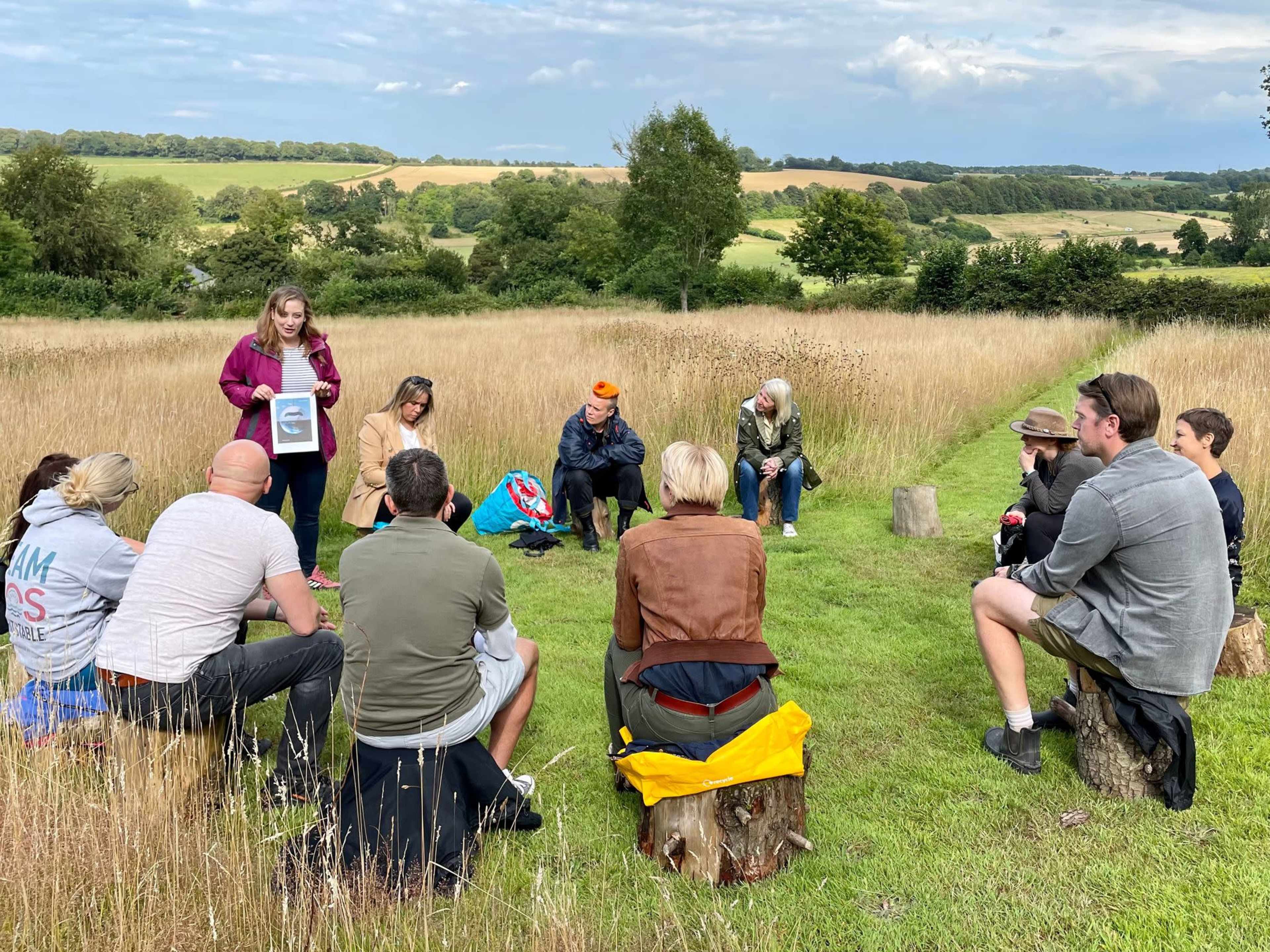 A group of people sits on logs in a grassy field while one person stands and holds a document, surrounded by trees and hills in the background.