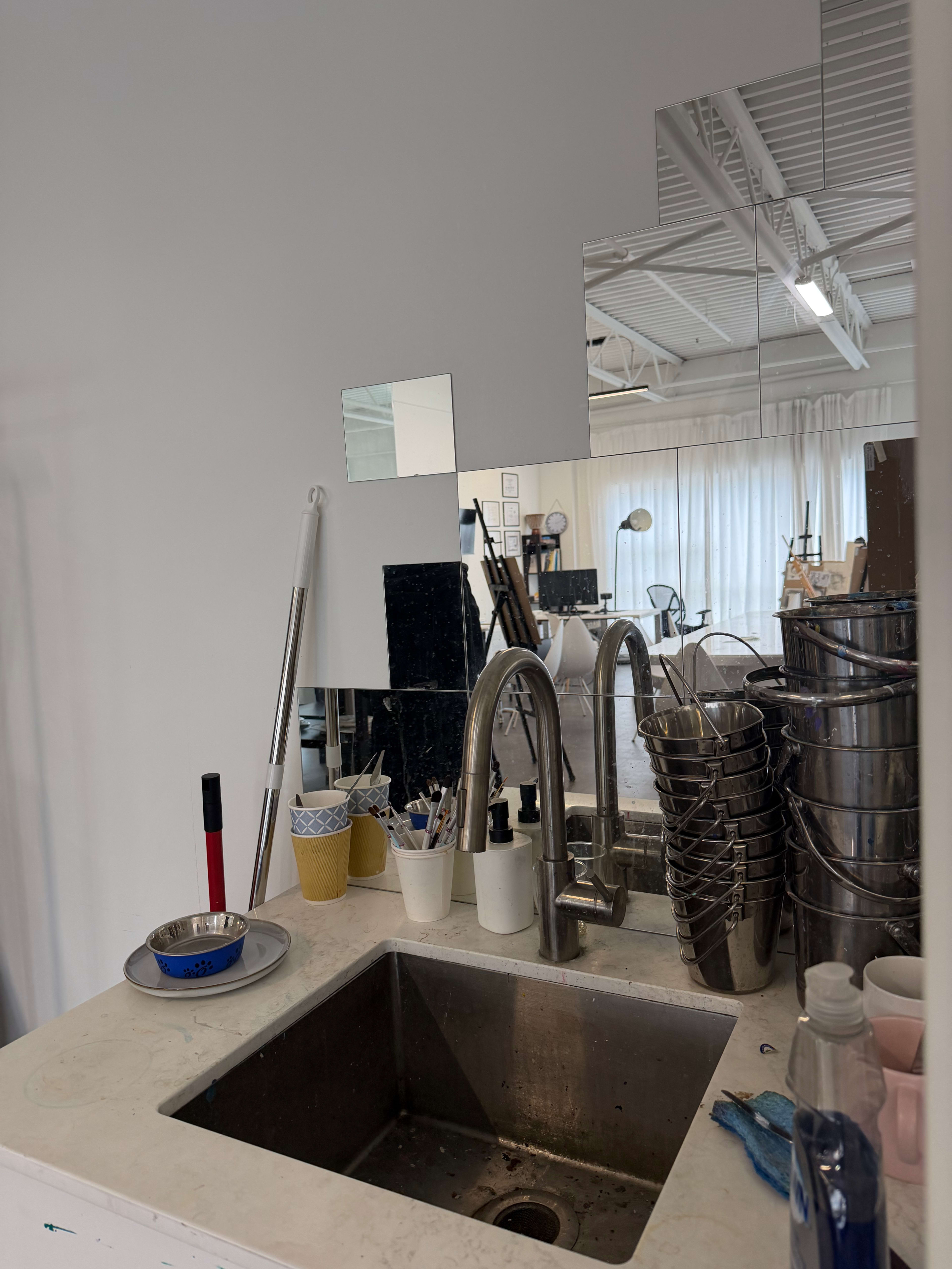 The image shows a stainless steel sink in a bright, spacious room with a mirror and various utensils organized around it.