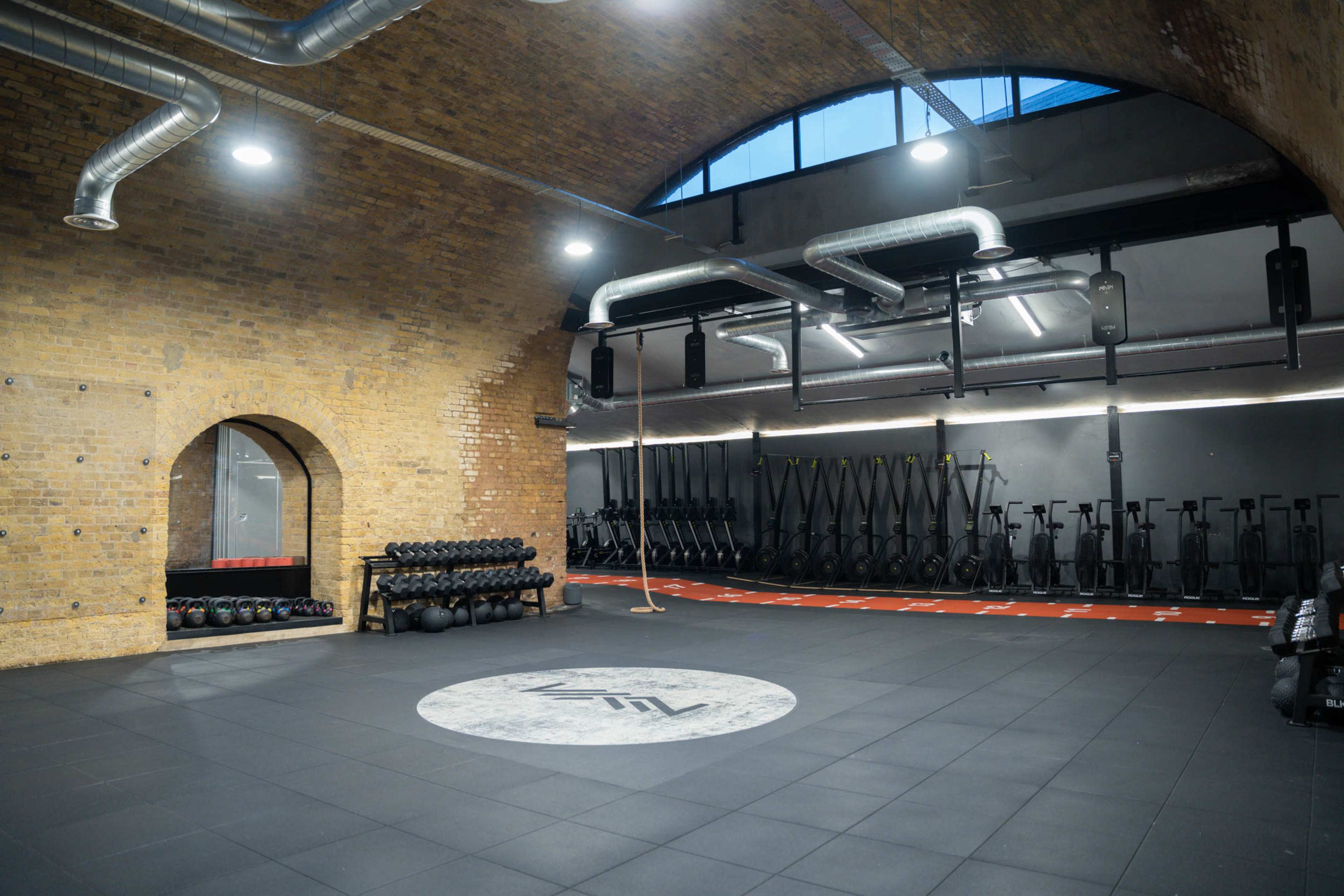 A spacious gym with exposed brick walls, featuring weights on shelves, exercise bikes, and a large circular logo on the floor.