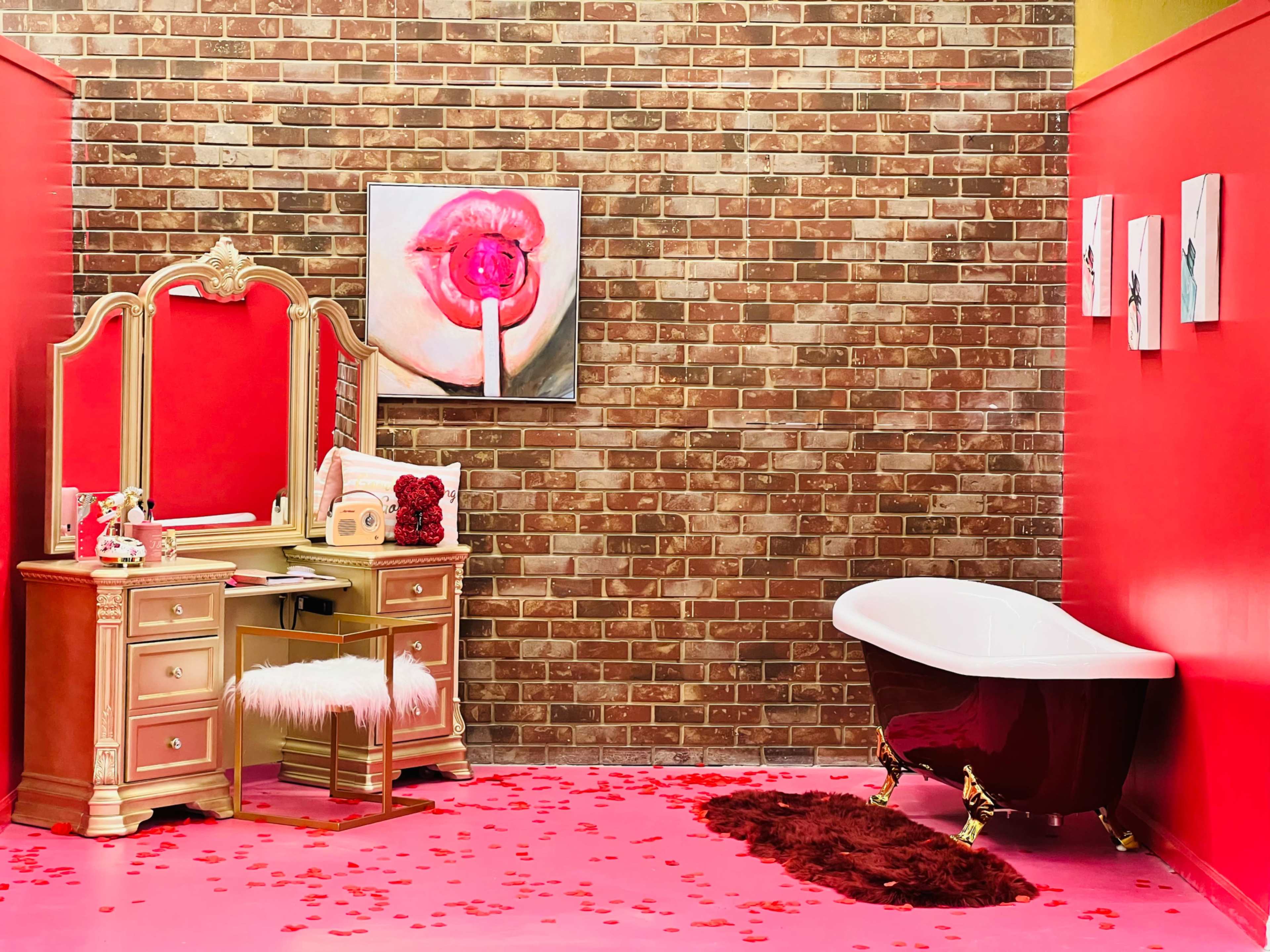 A bathroom featuring a brick wall, a red-painted space with a vintage vanity, a white freestanding bathtub, and a pink carpet scattered with rose petals.