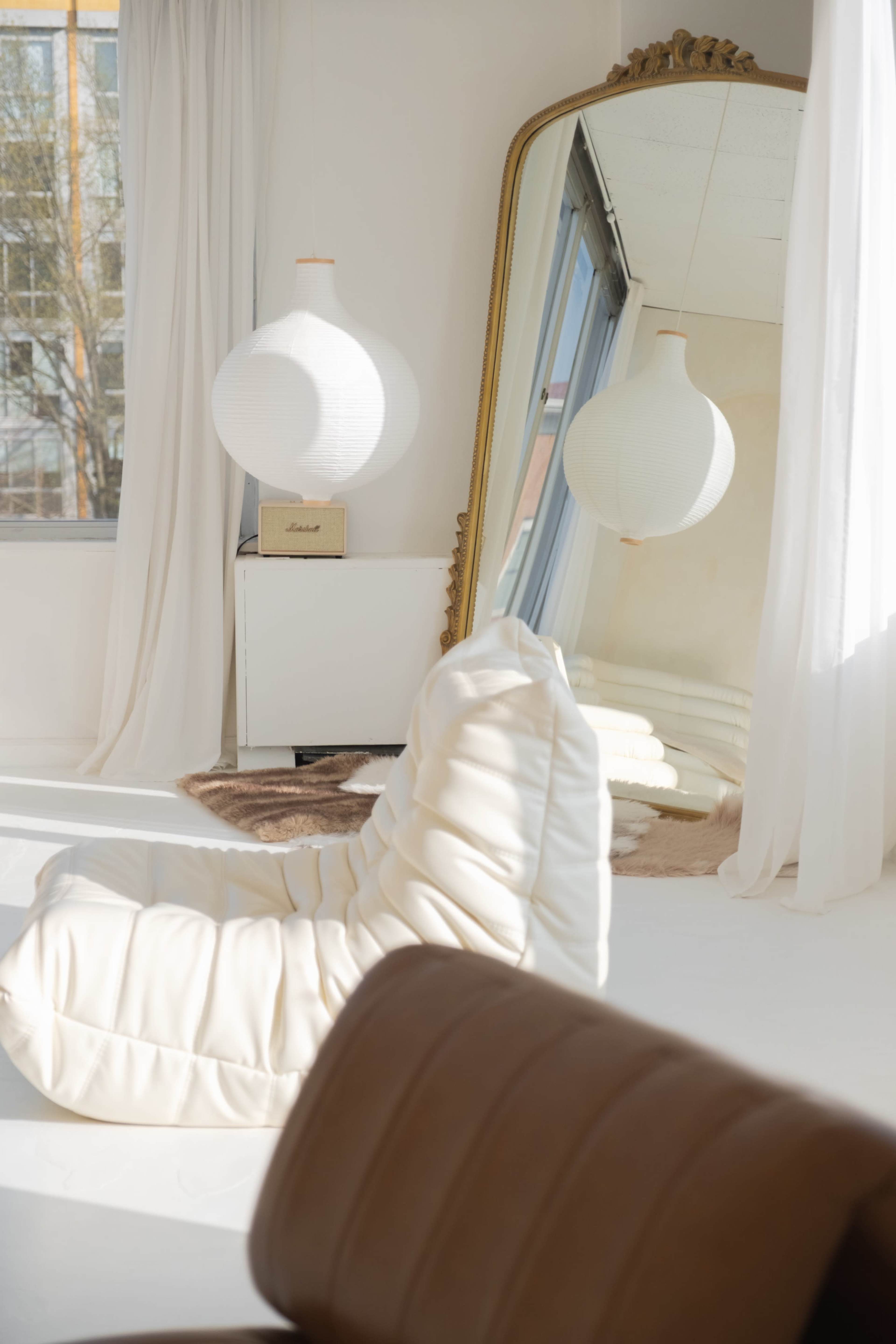 The image shows a bright, minimalist room with a large mirror, a white floor lamp, and a cream-colored bean bag chair.