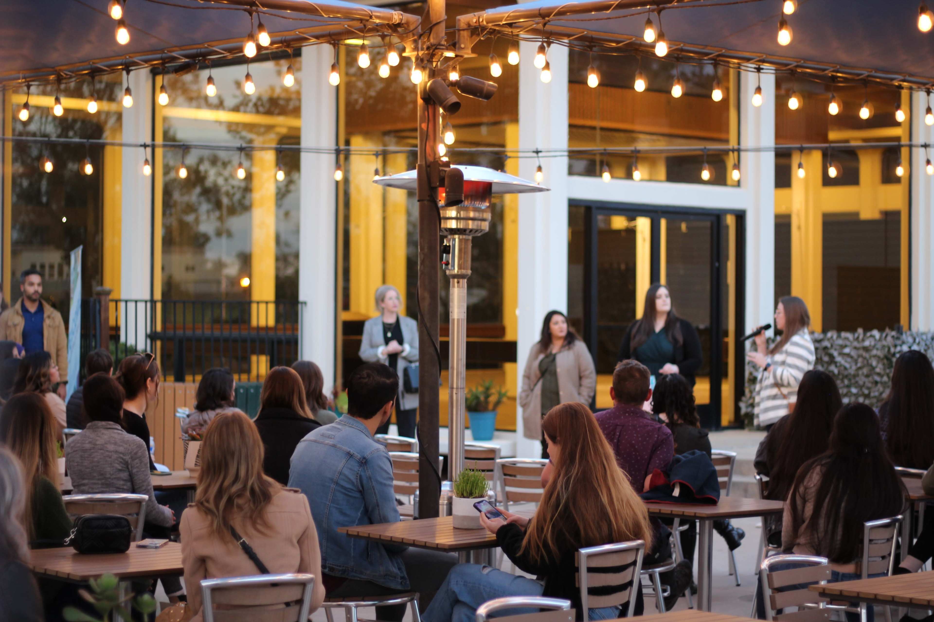 A group of people listens to speakers in an outdoor seating area illuminated by string lights.