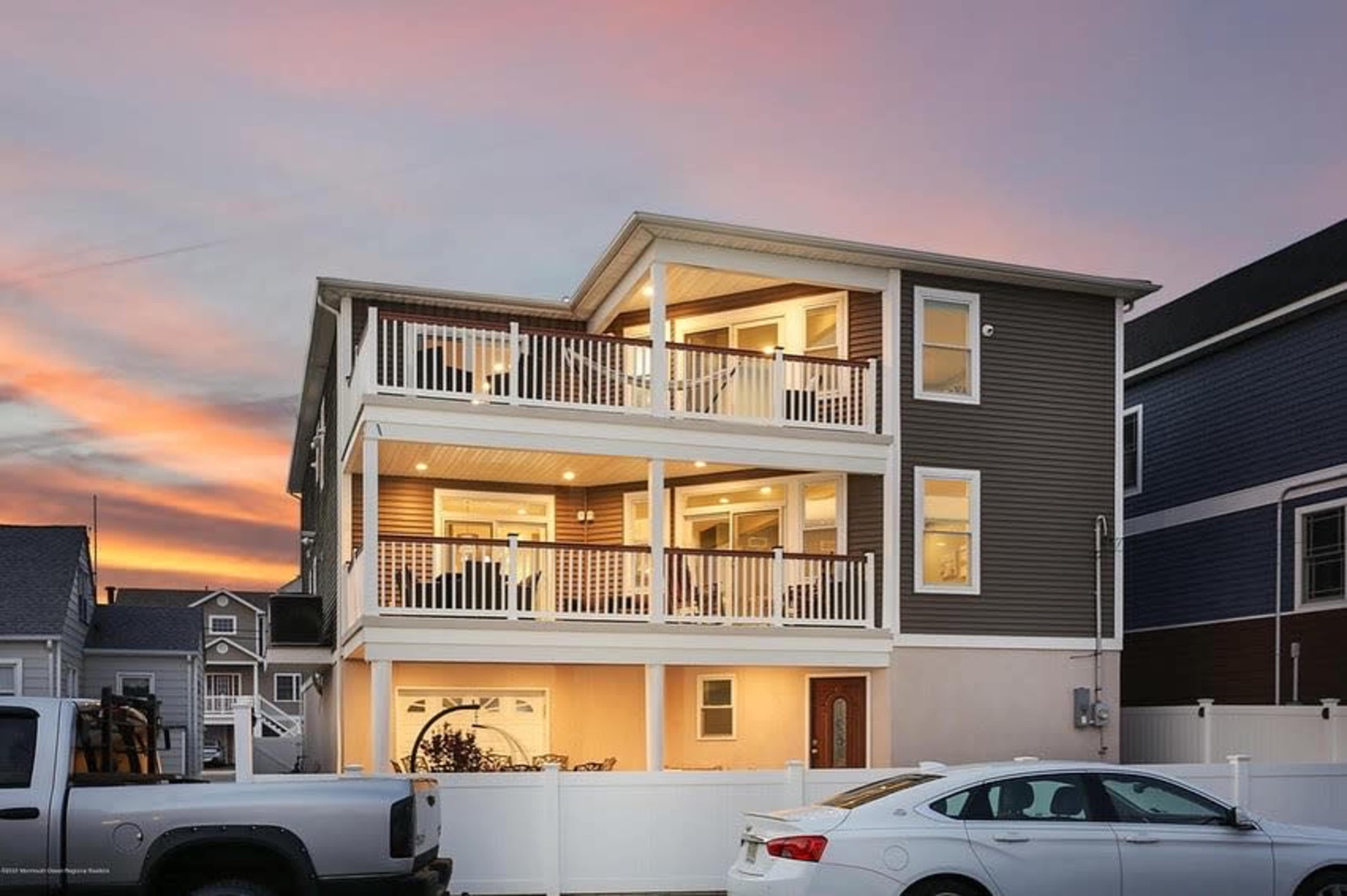 A three-story house features two balconies with white railings and large windows, illuminated at sunset, surrounded by a white fence and parked cars.