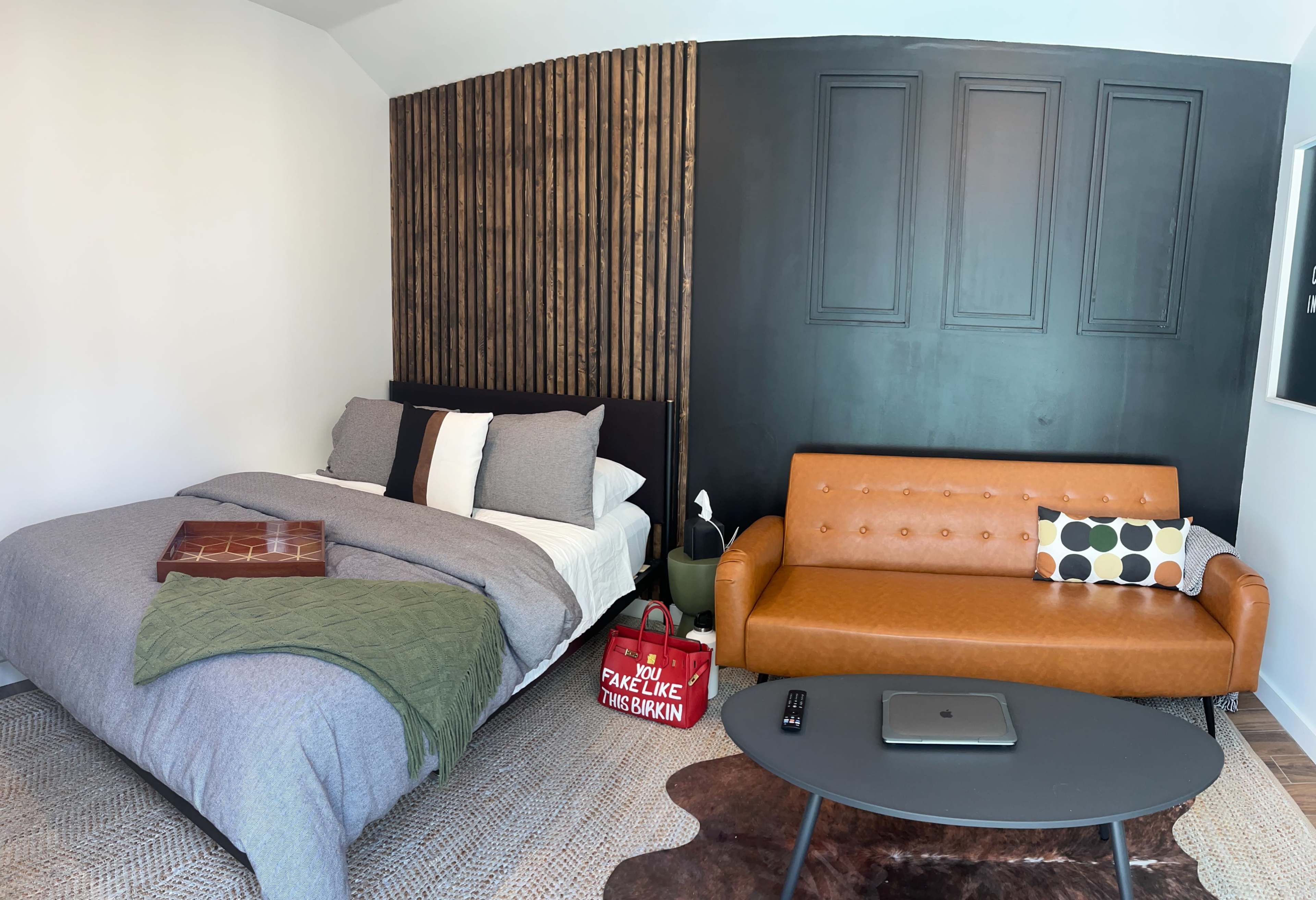 A minimalist bedroom featuring a bed with gray bedding, a brown leather sofa, and a small round table in front.