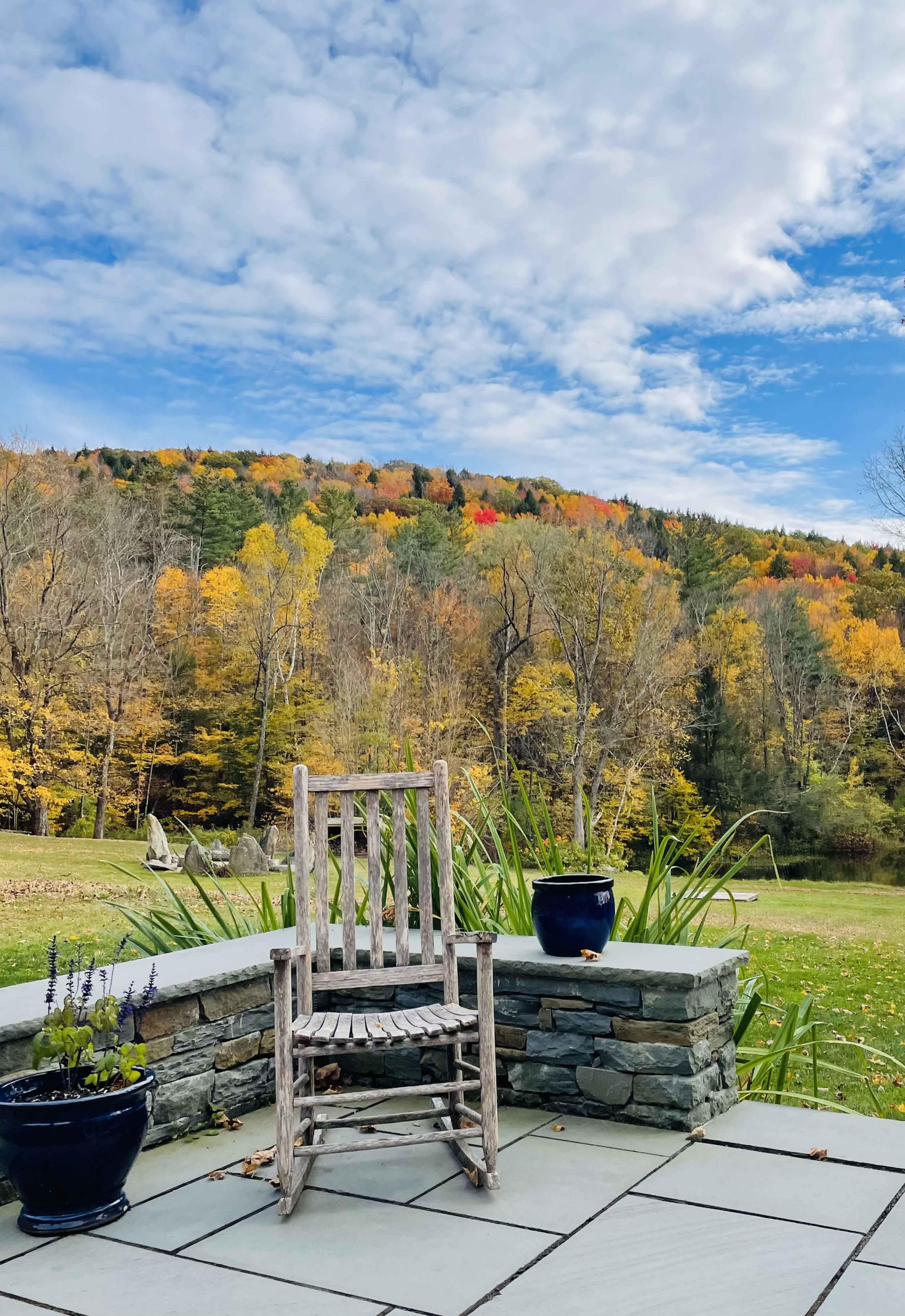 A rustic wooden rocking chair sits on a stone patio surrounded by colorful autumn foliage and potted plants.