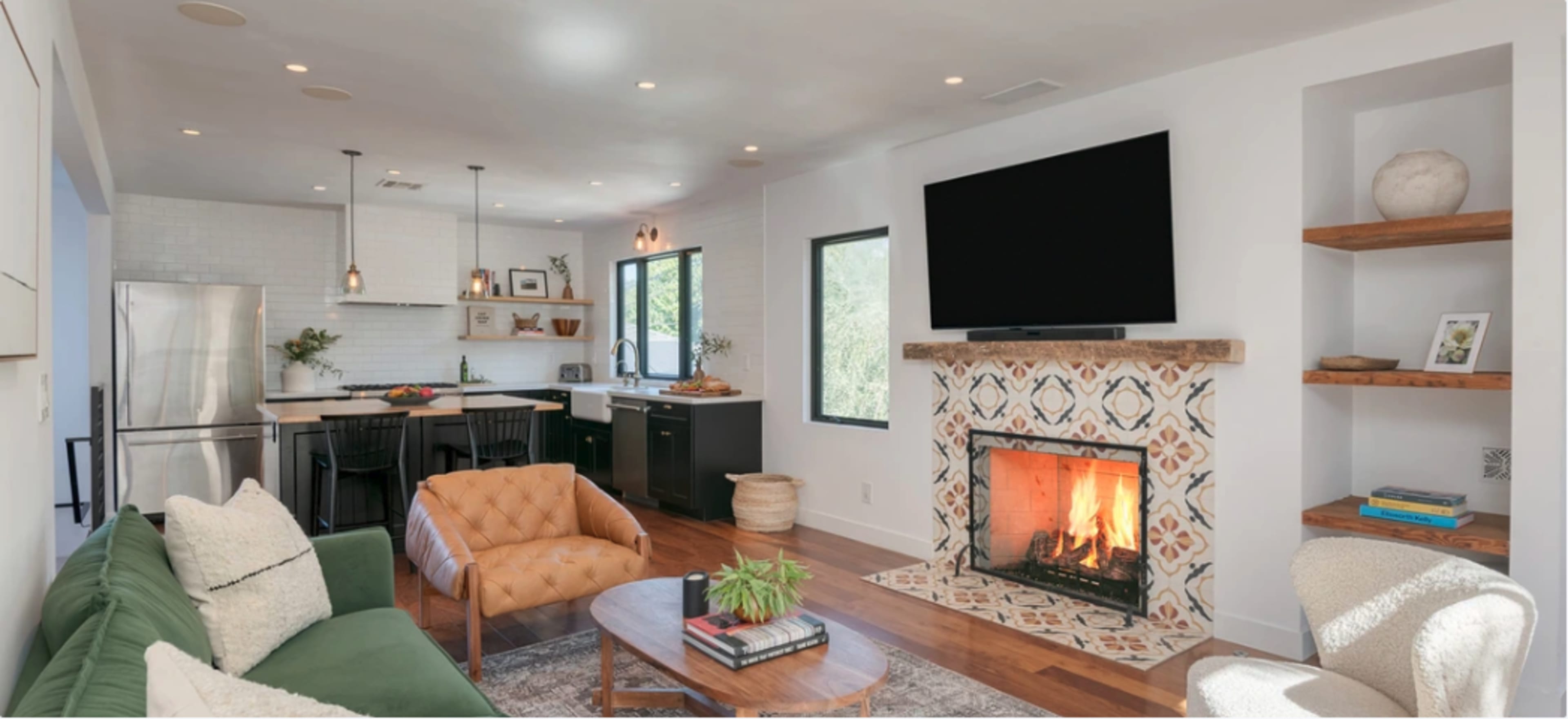 A modern living room featuring a green sofa, an orange leather armchair, a wooden coffee table, and a fireplace with decorative tiles, all in a space connected to a kitchen with black cabinets and white countertops.