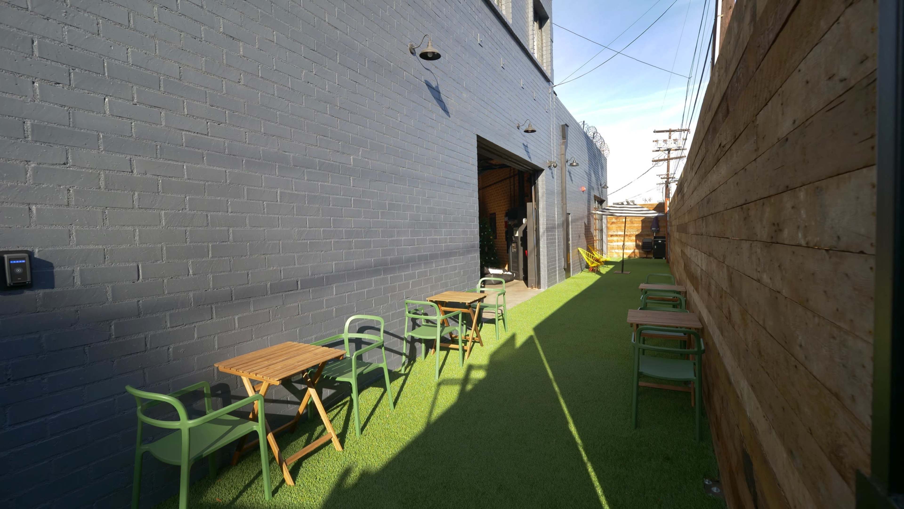 The image shows a narrow outdoor space with green artificial grass, wooden tables, and chairs along a brick wall and an open entrance to a building.