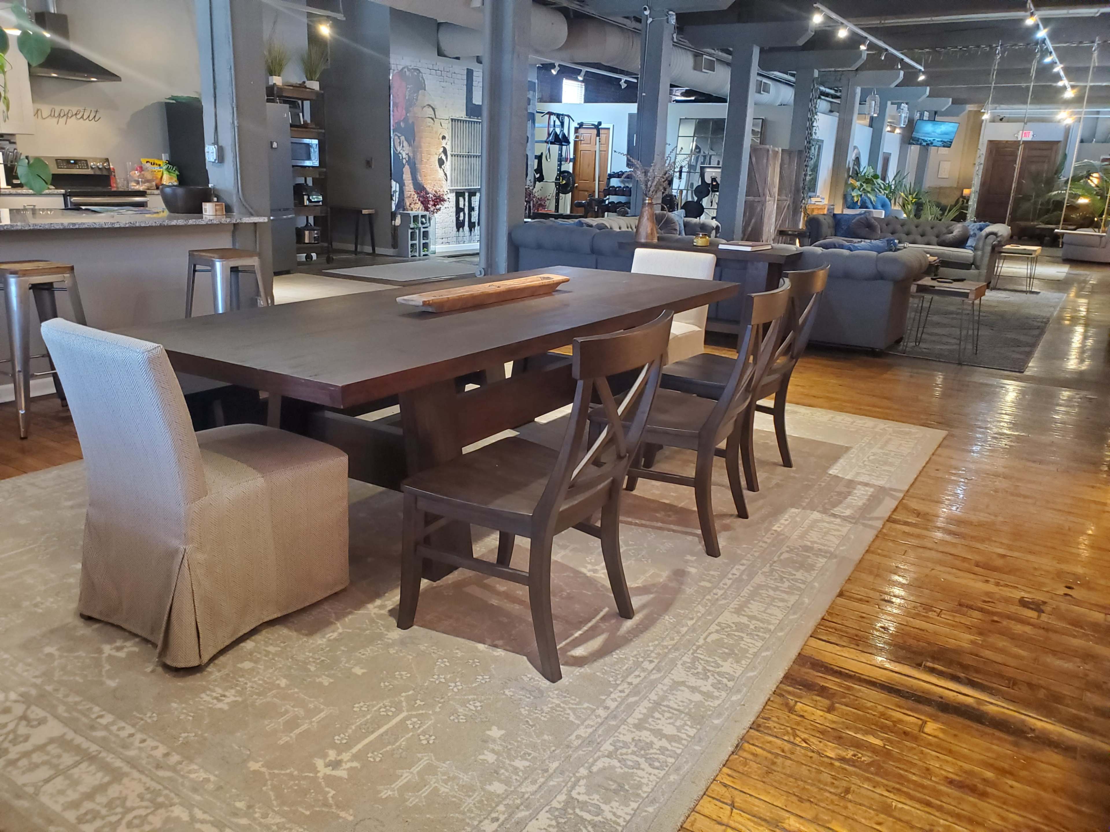 A large wooden dining table with six chairs is set on a patterned rug in a spacious room filled with modern furniture and decor.