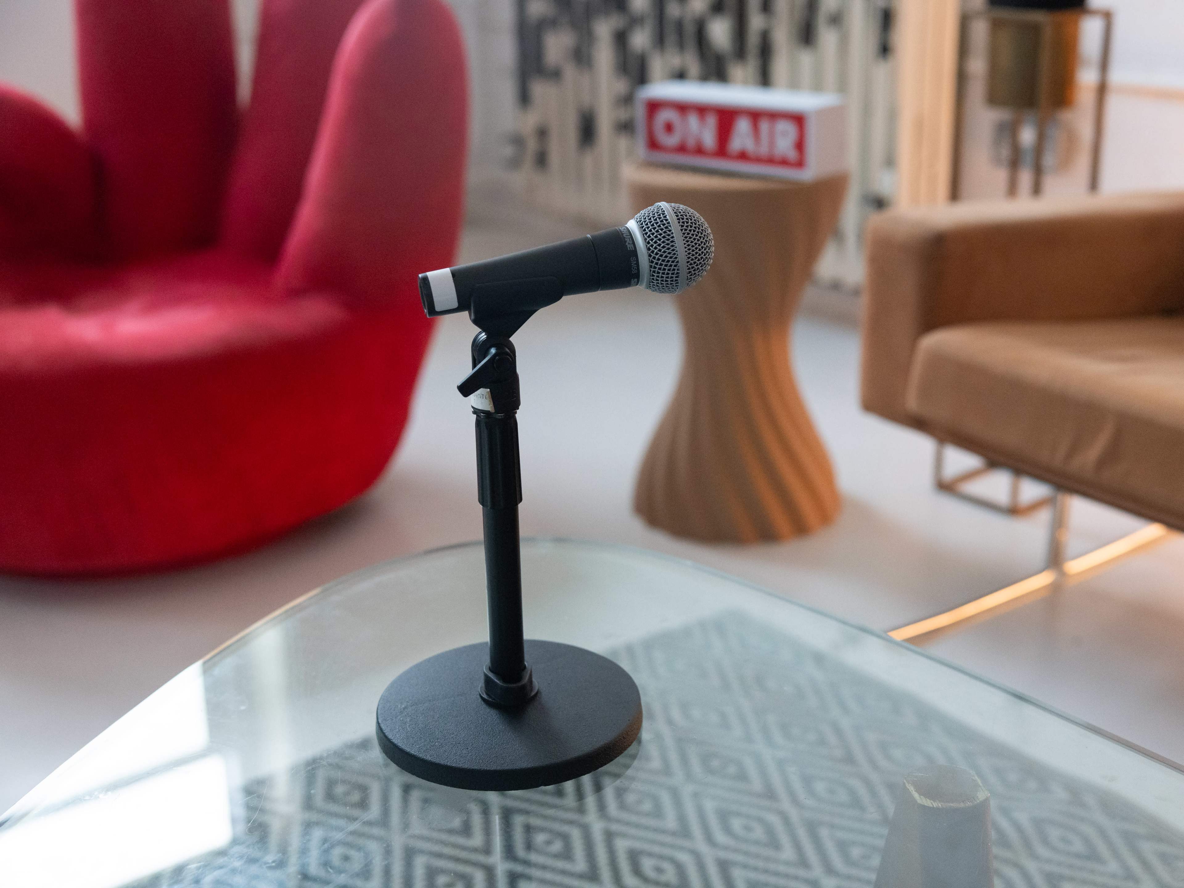 A black microphone stands on a circular base in front of a glass table, with a red chair and a beige sofa visible in the background.