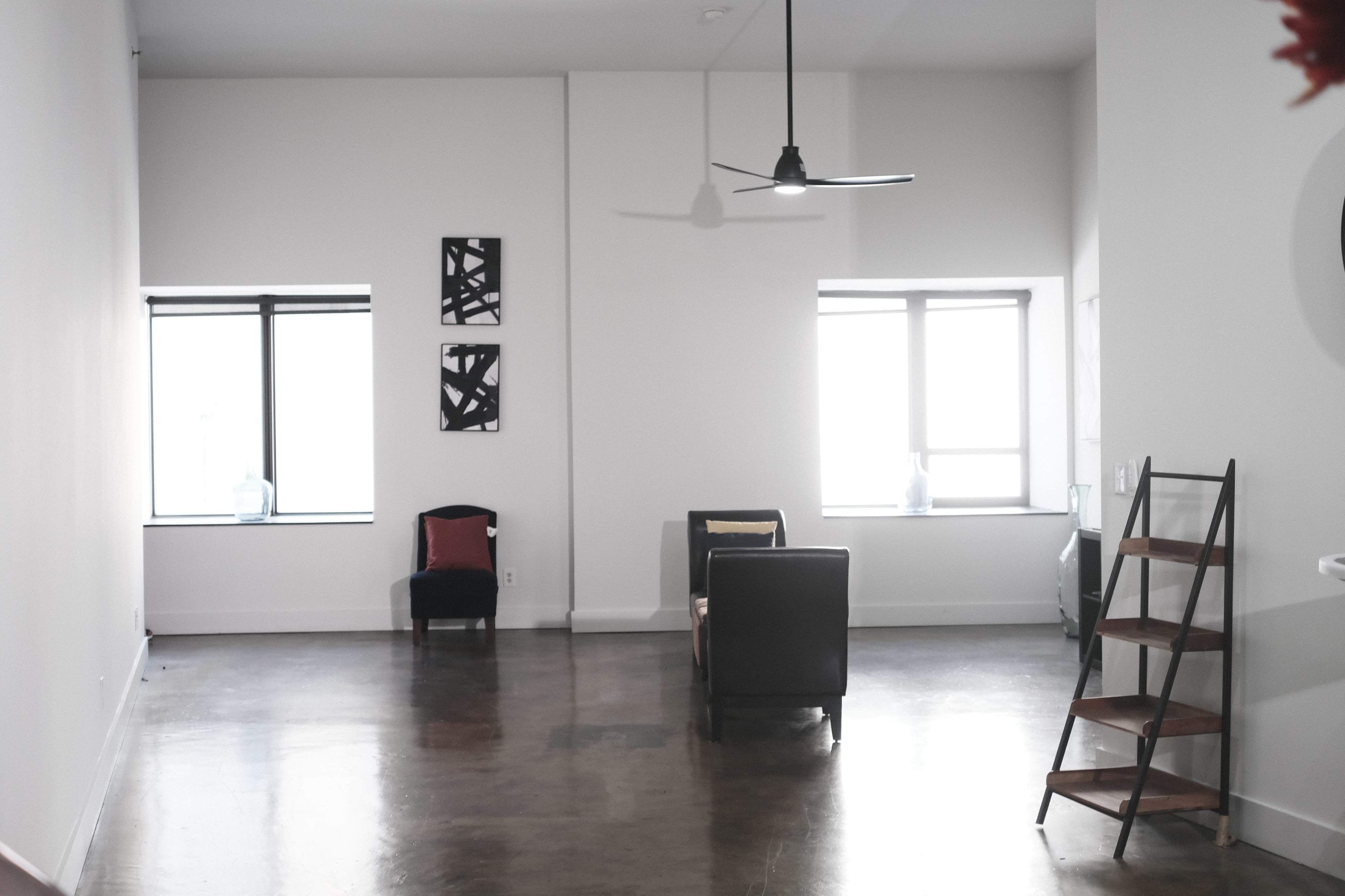 An empty room features a chair, a sofa, and a ladder shelf against white walls with two windows allowing light to enter.