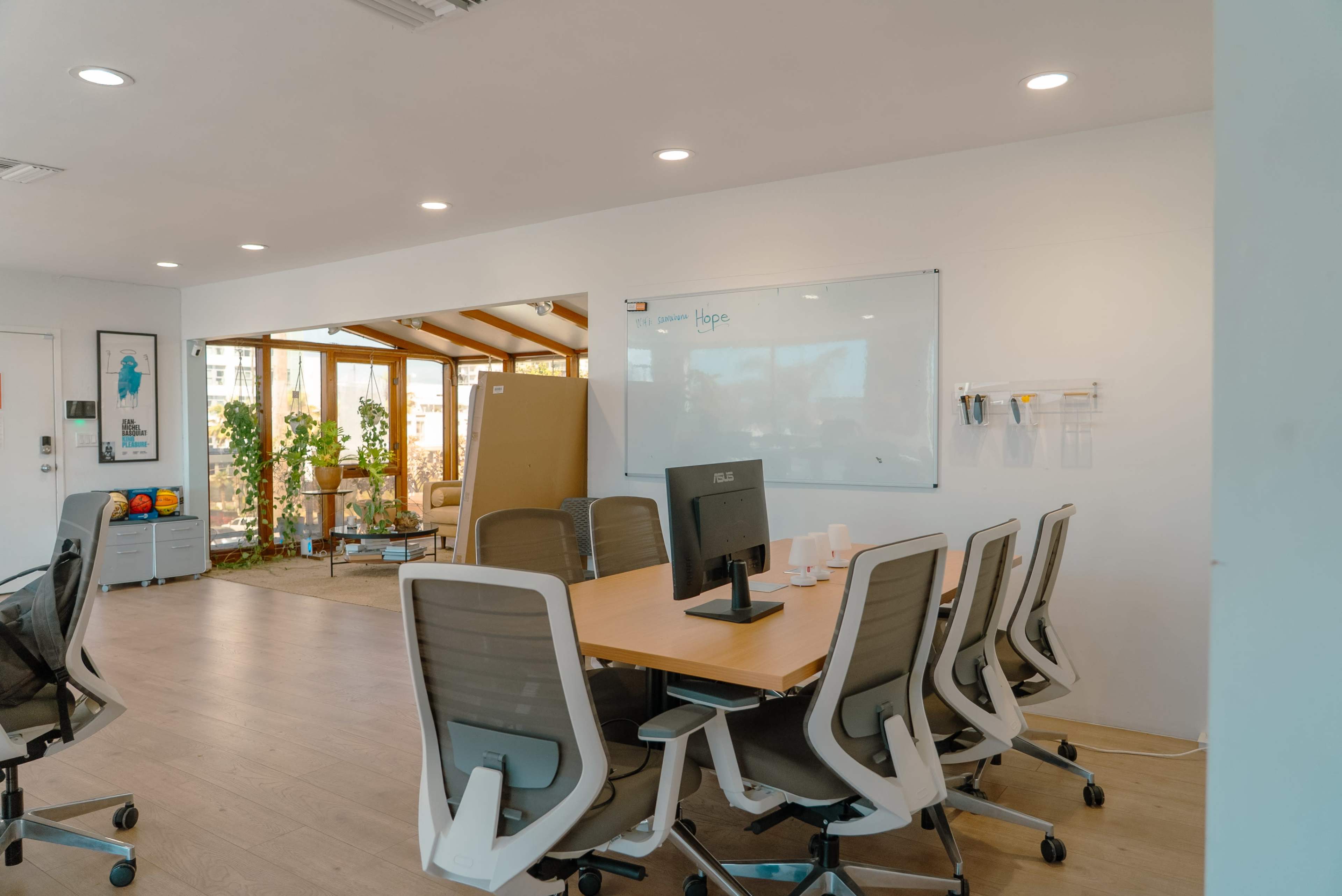The image shows a modern office space featuring a conference table surrounded by ergonomic chairs and a whiteboard on the wall, with a sunny area containing plants visible in the background.