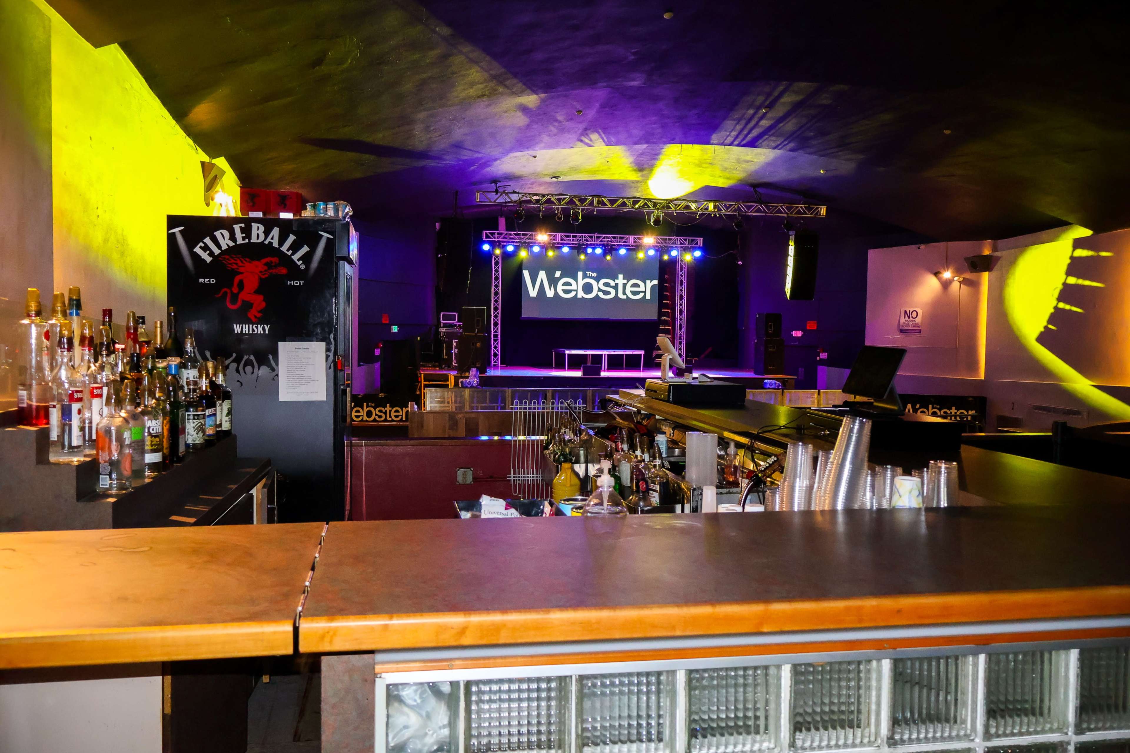 The interior of a music venue featuring a bar area with a variety of liquor bottles, and a stage in the background illuminated with colorful lights.