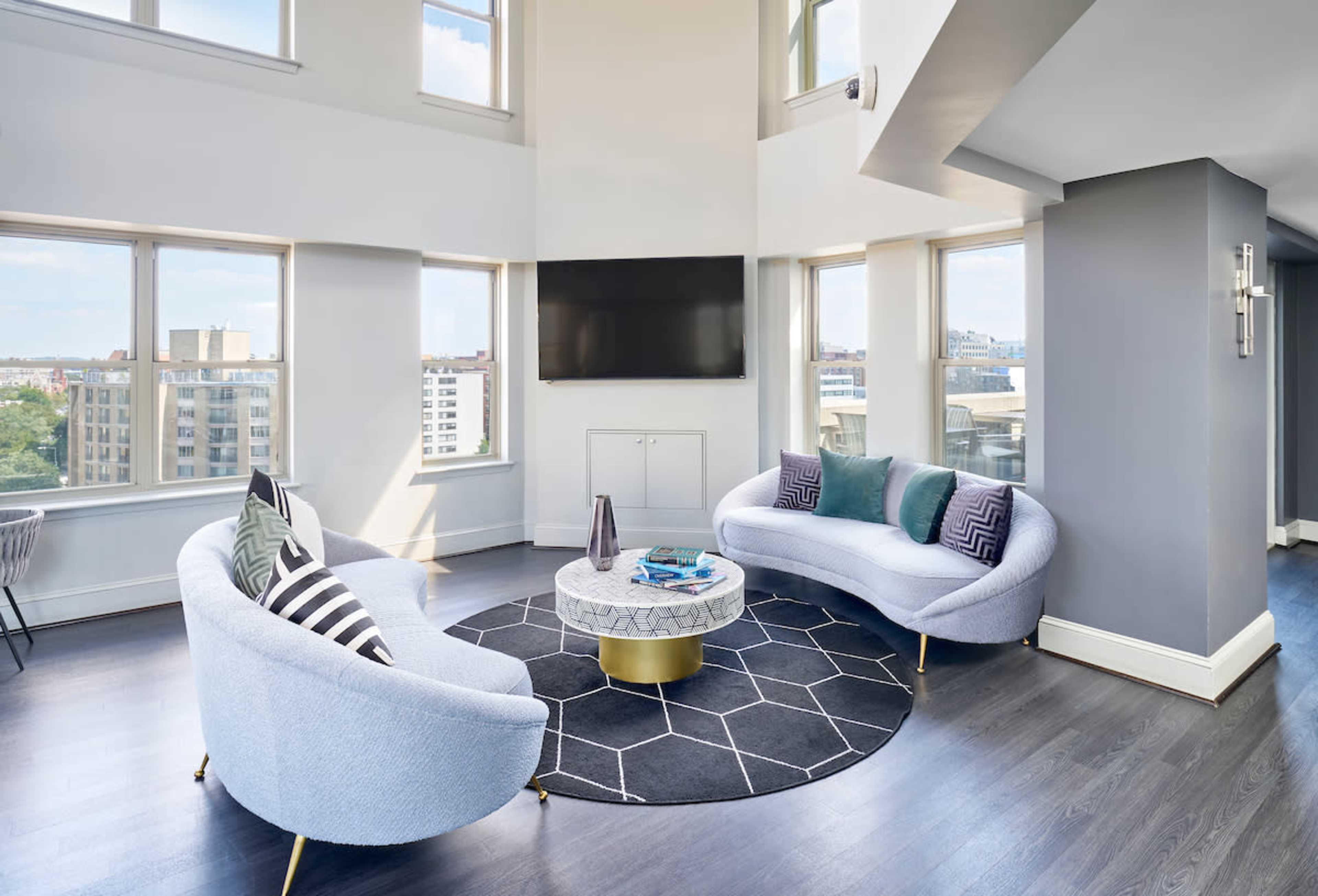 A bright, modern living area features two curved sofas, a circular coffee table, and large windows providing a view of the city.