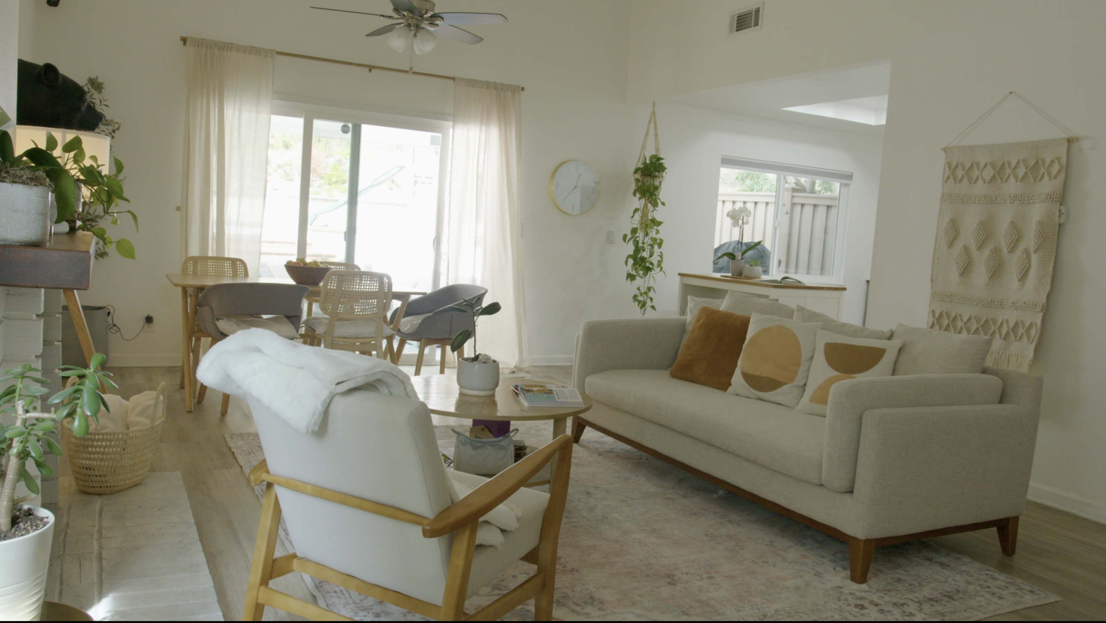 A bright living room features a sofa with decorative pillows, a wooden coffee table, and a dining area with chairs near large windows.