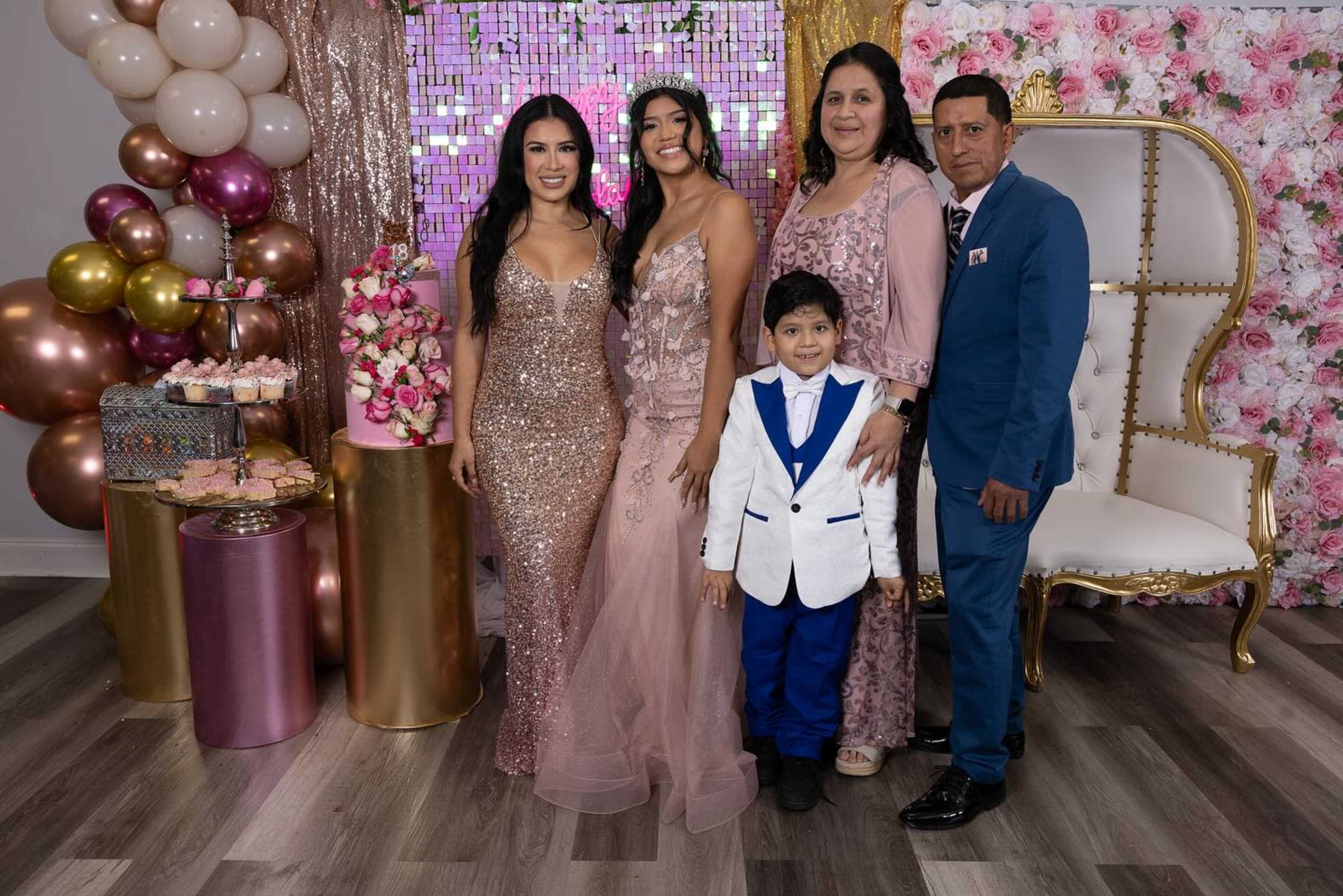 A group of five people poses together in a decorated room featuring floral backdrops and balloon arrangements for a celebration.