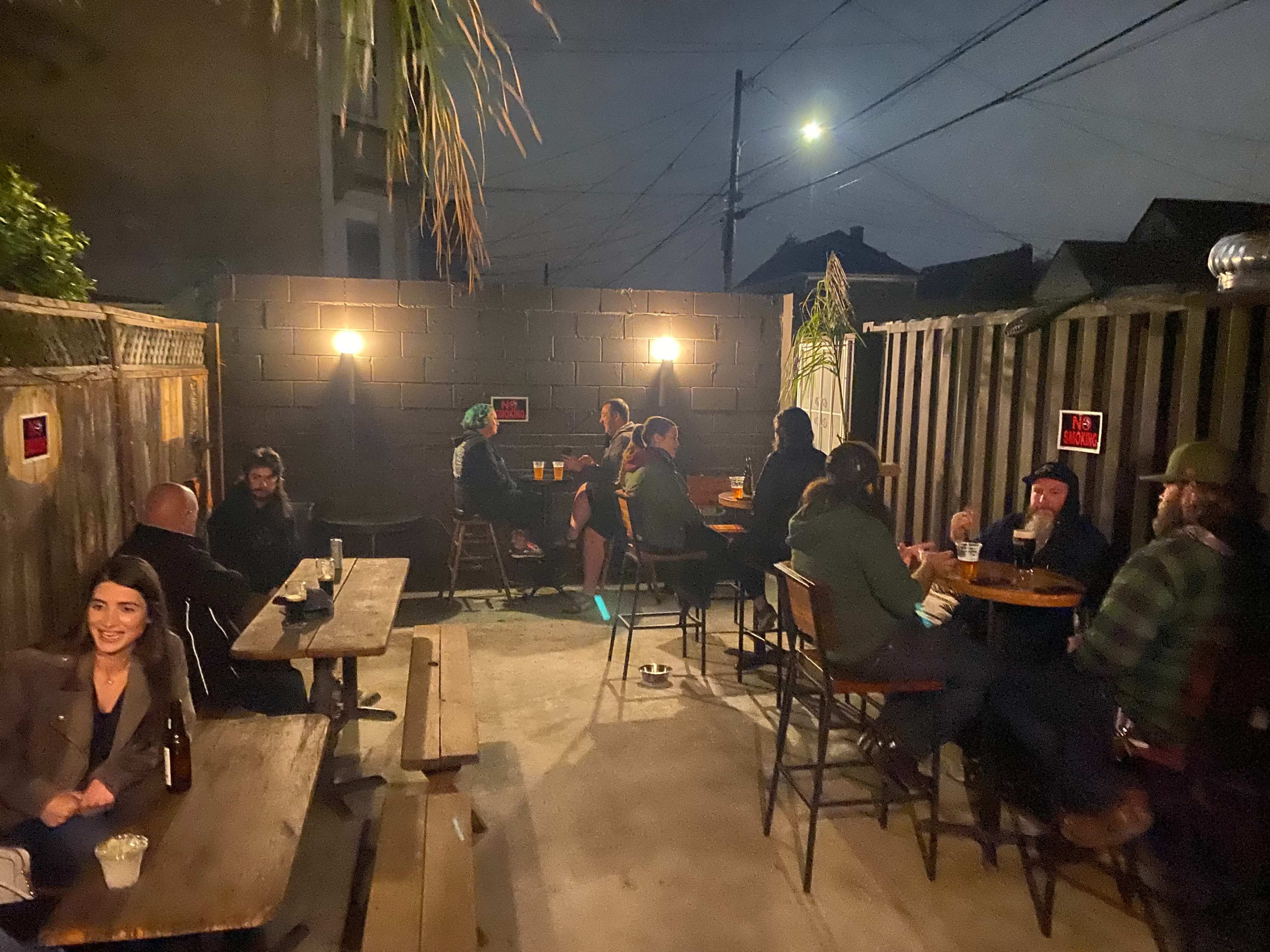 A group of people sits at wooden tables in an outdoor space at night, surrounded by low walls and illuminated by soft lights.