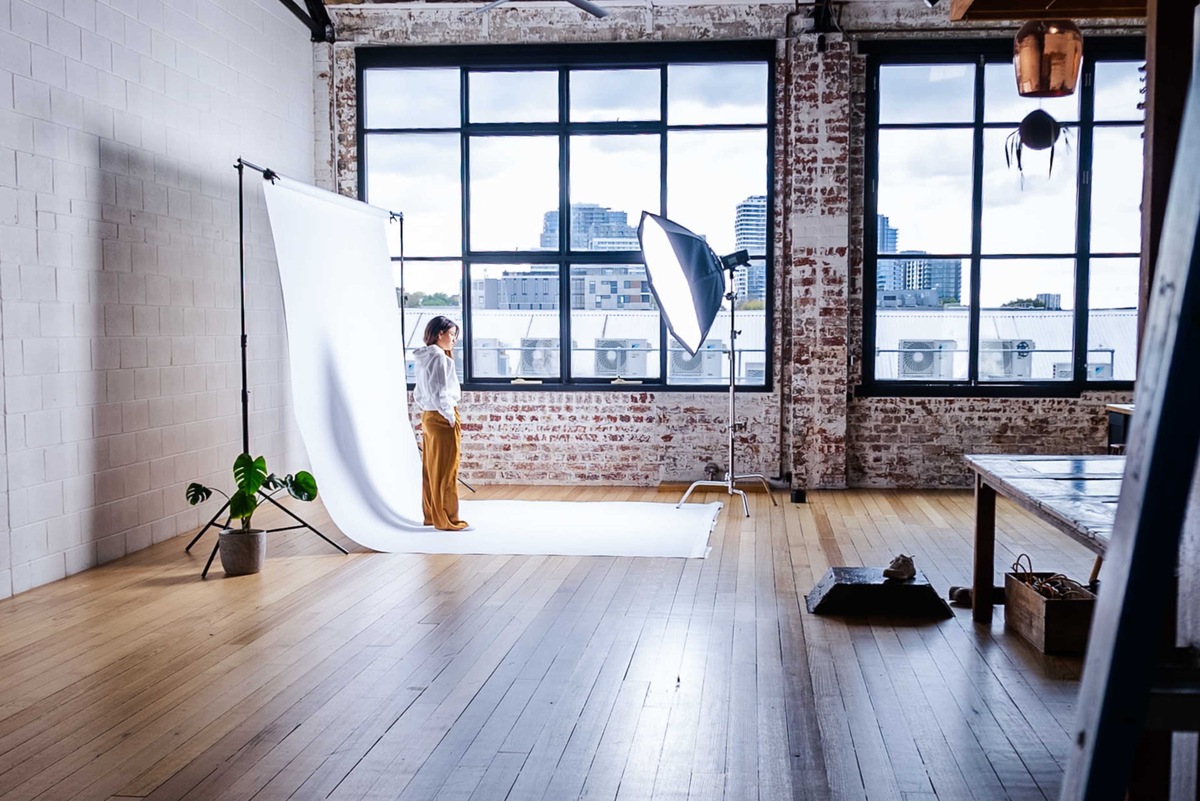 A person stands on a white backdrop in a spacious studio with large windows and exposed brick walls.