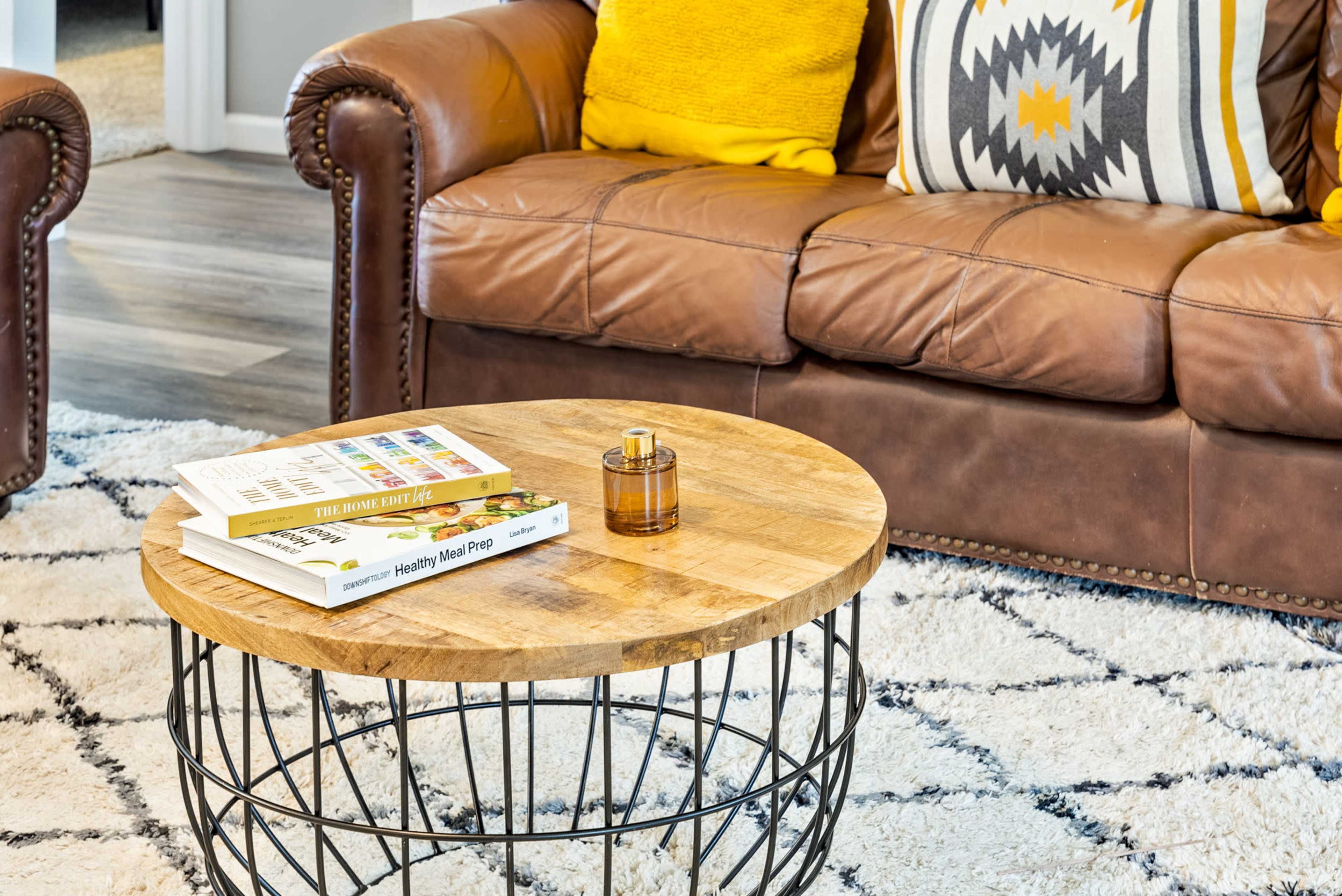 A leather sofa with decorative pillows sits next to a wooden coffee table, which holds books and a small glass candle.