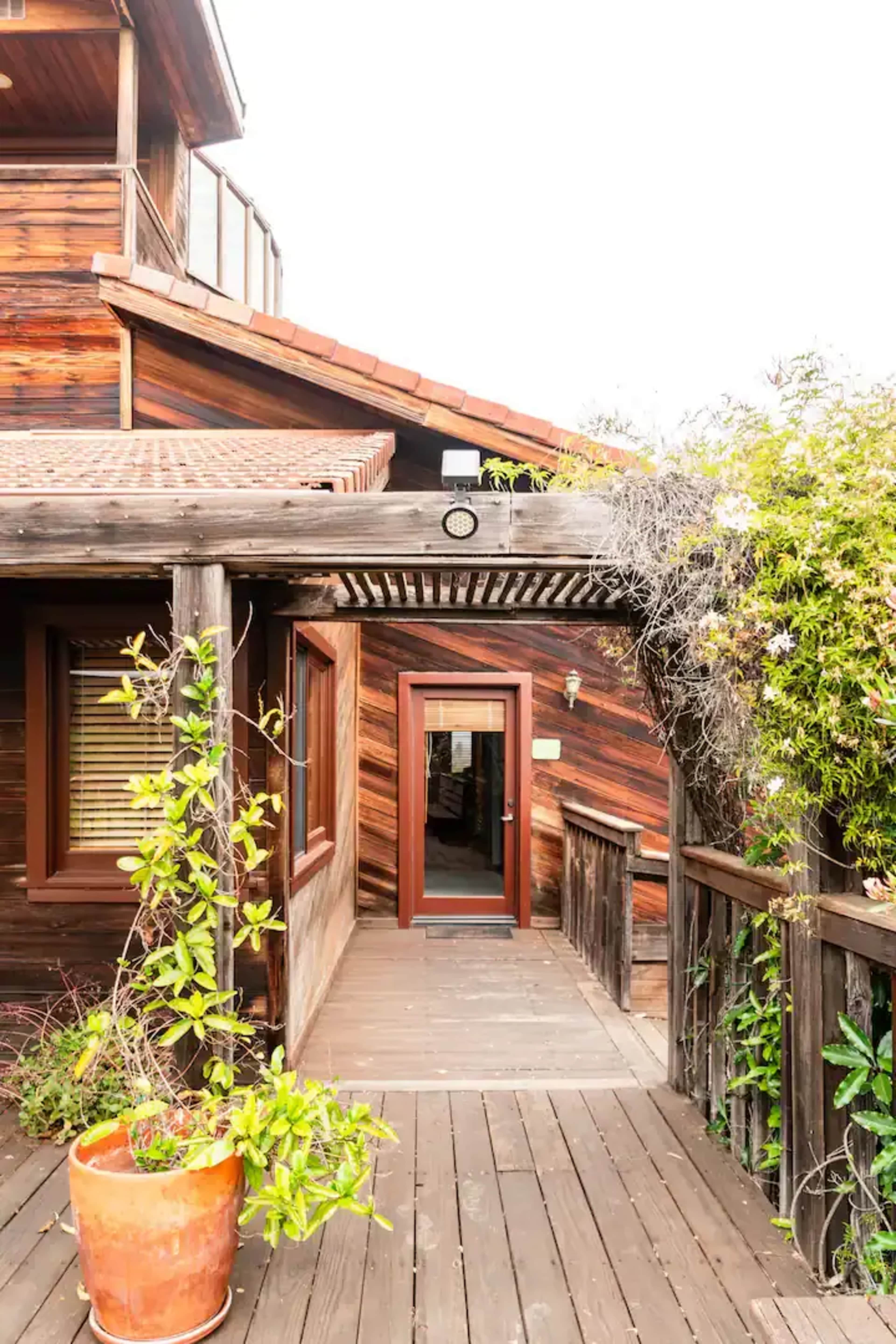 The image shows a wooden deck leading to a doorway of a house surrounded by greenery and a potted plant.