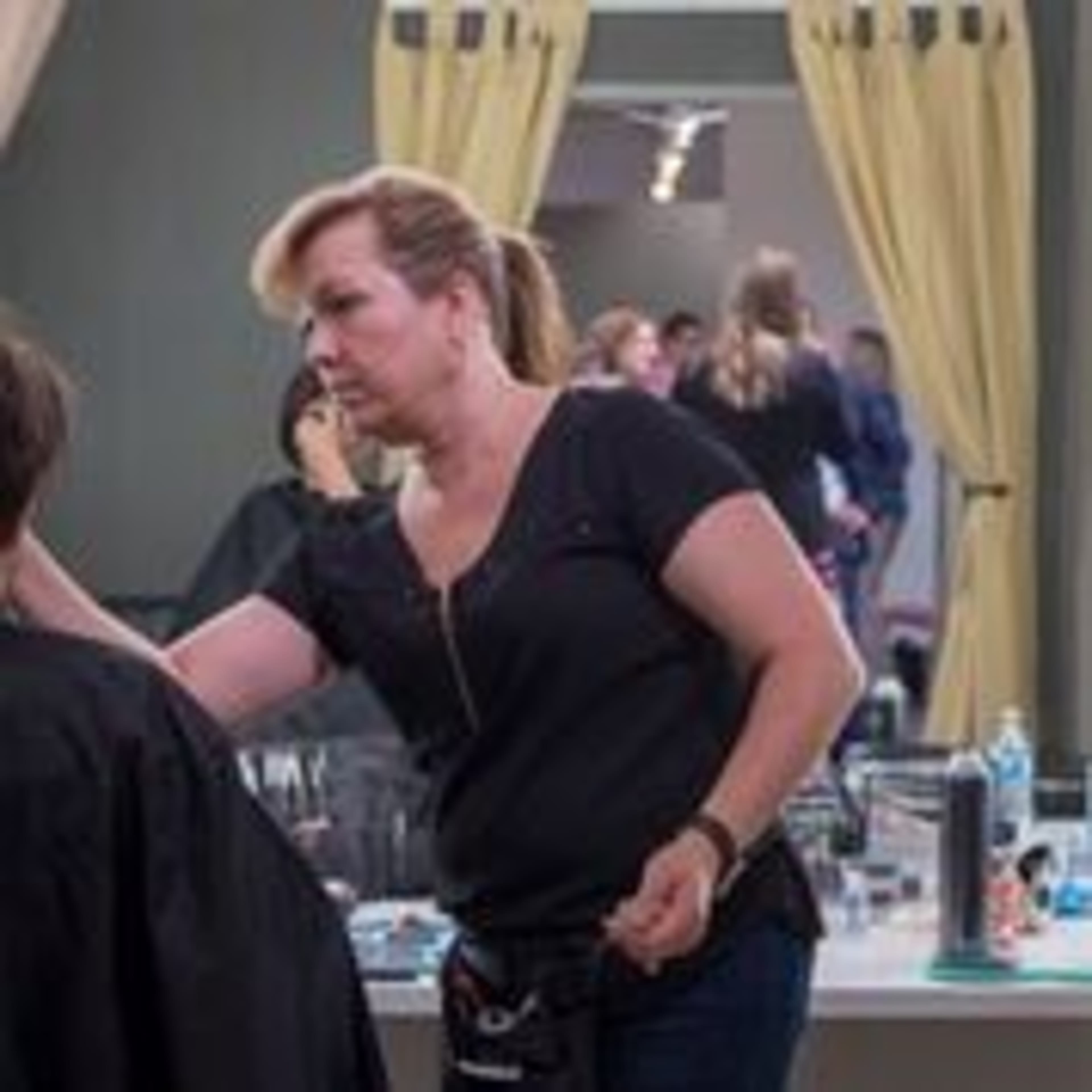 A hairstylist works on a client's hair at a salon, with other patrons visible in the background.