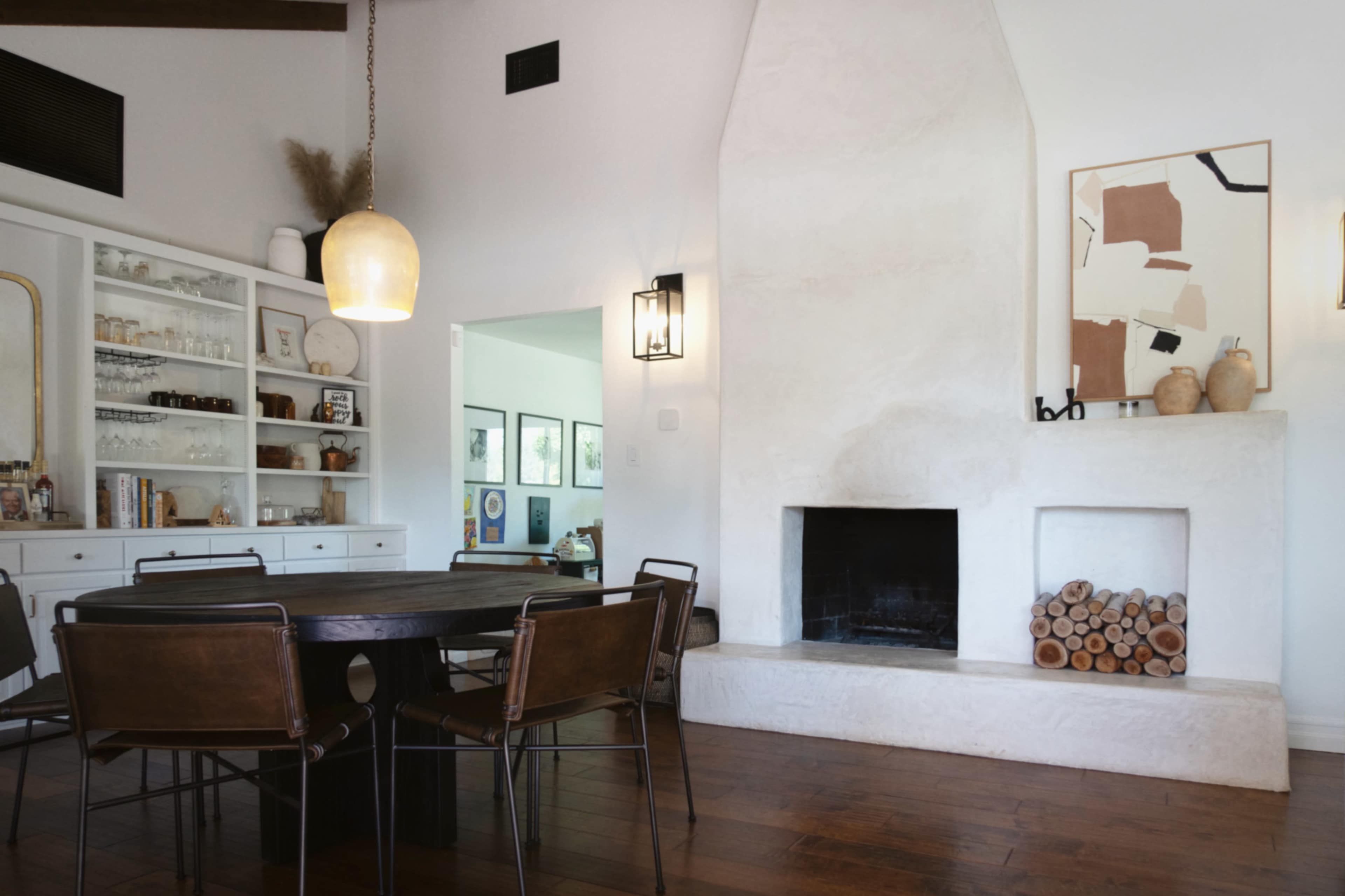 A dining area features a round wooden table surrounded by black metal chairs, with a white plaster fireplace and a collection of stacked firewood against the wall.