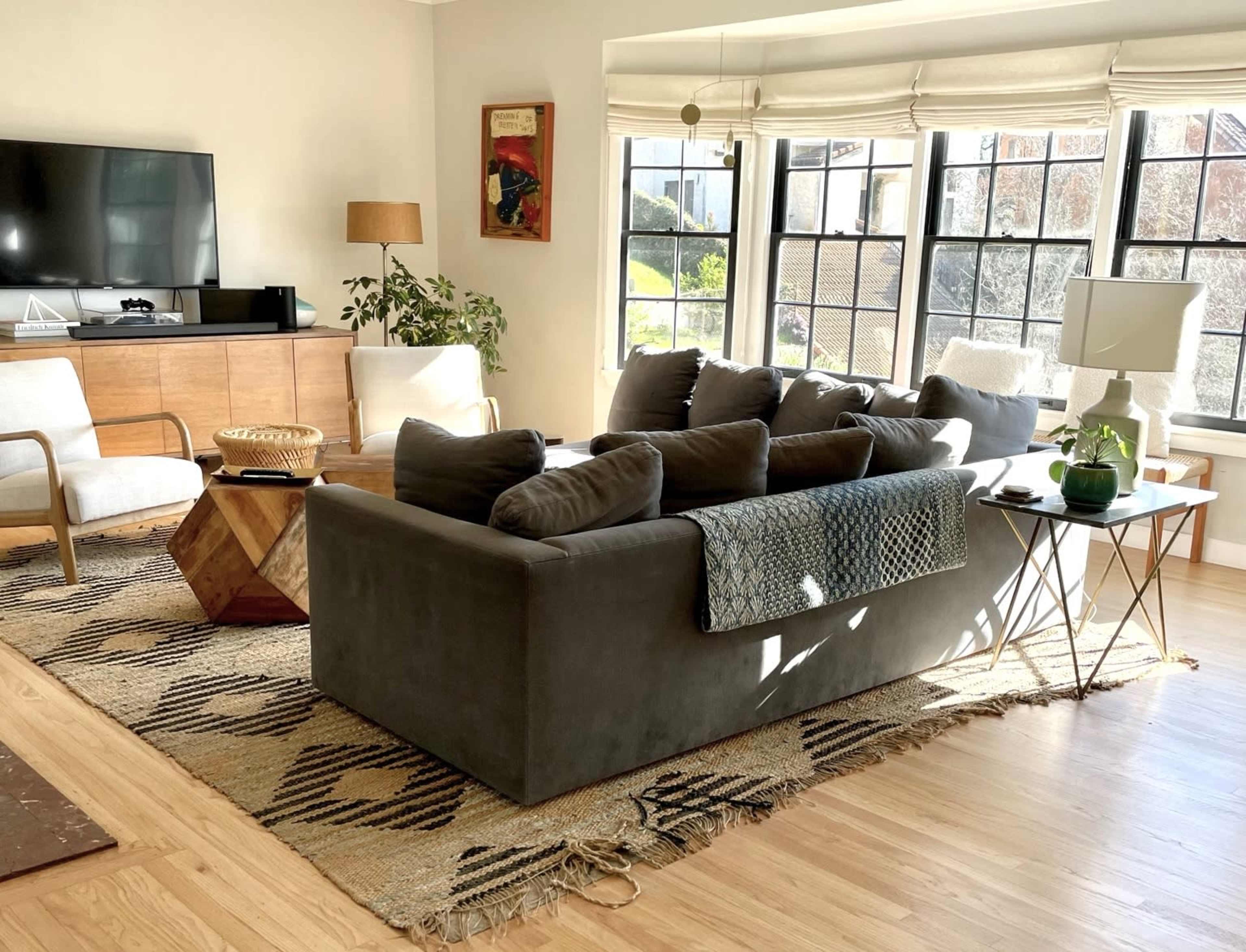 The living room features a gray sectional sofa with multiple cushions, a wooden coffee table, and large windows allowing natural light to fill the space.