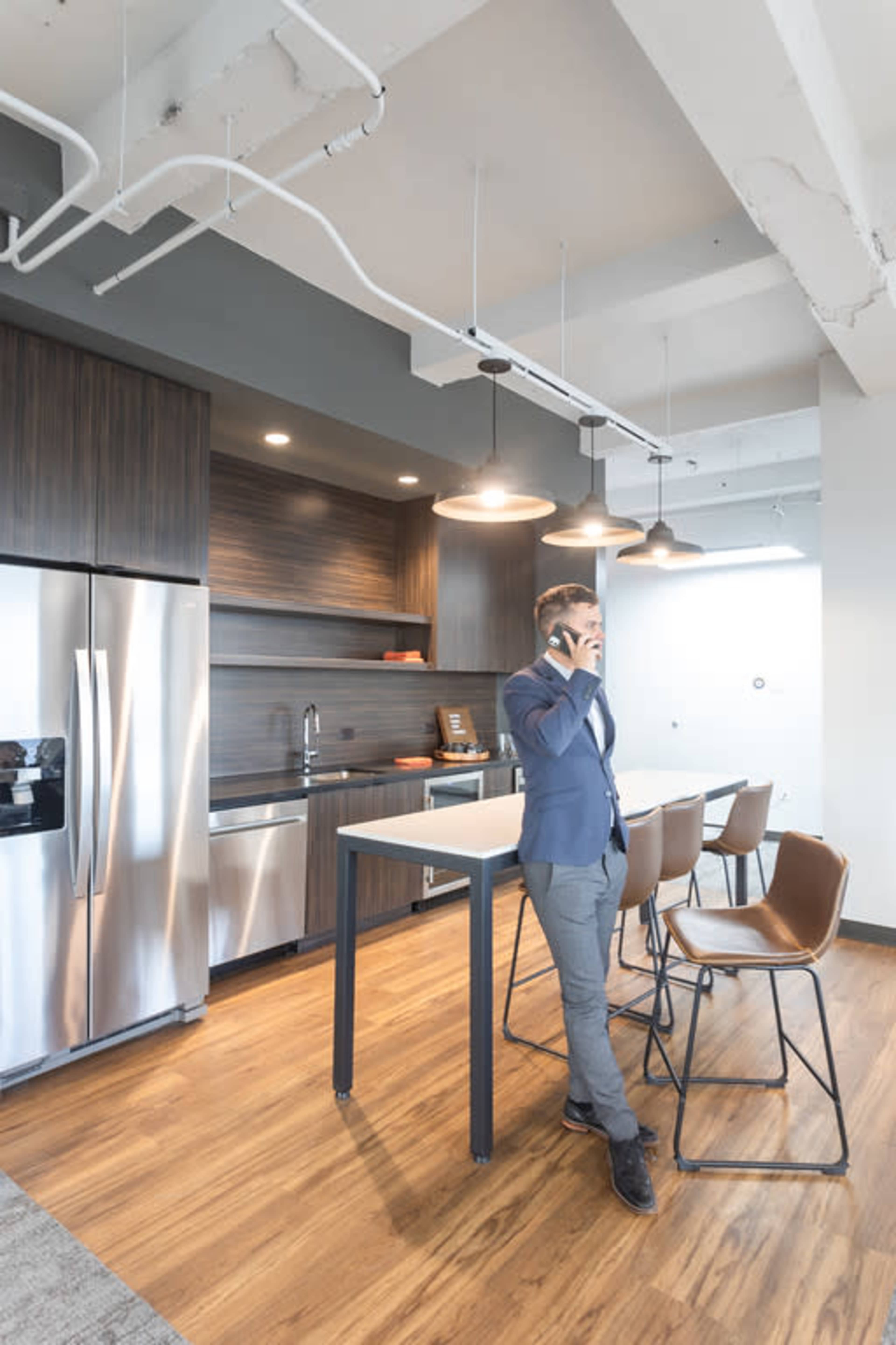 A man in a suit is standing by a modern kitchen with stainless steel appliances and a bar-style table.