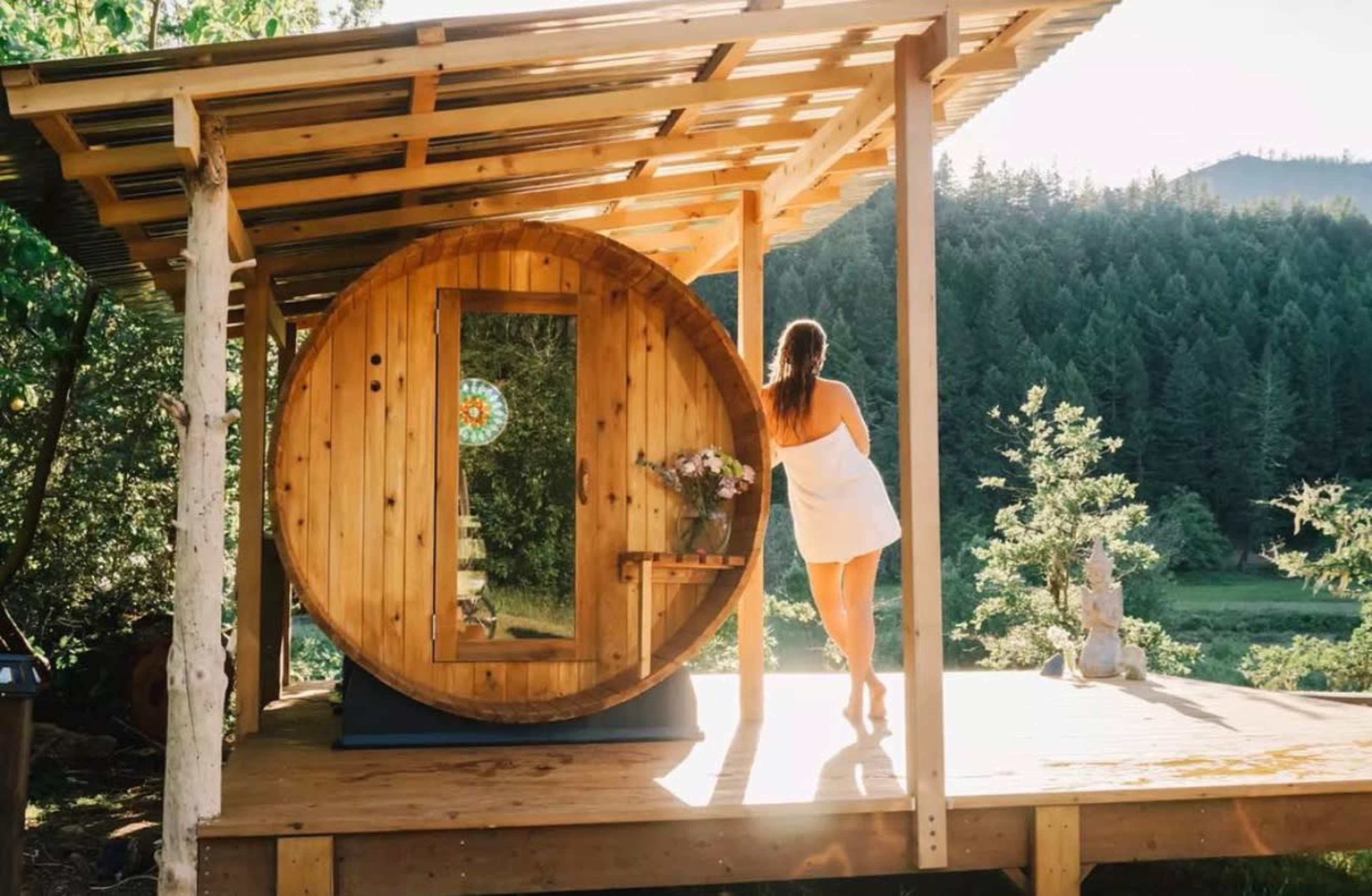 A woman stands on a deck next to a round wooden sauna, surrounded by trees and mountains.
