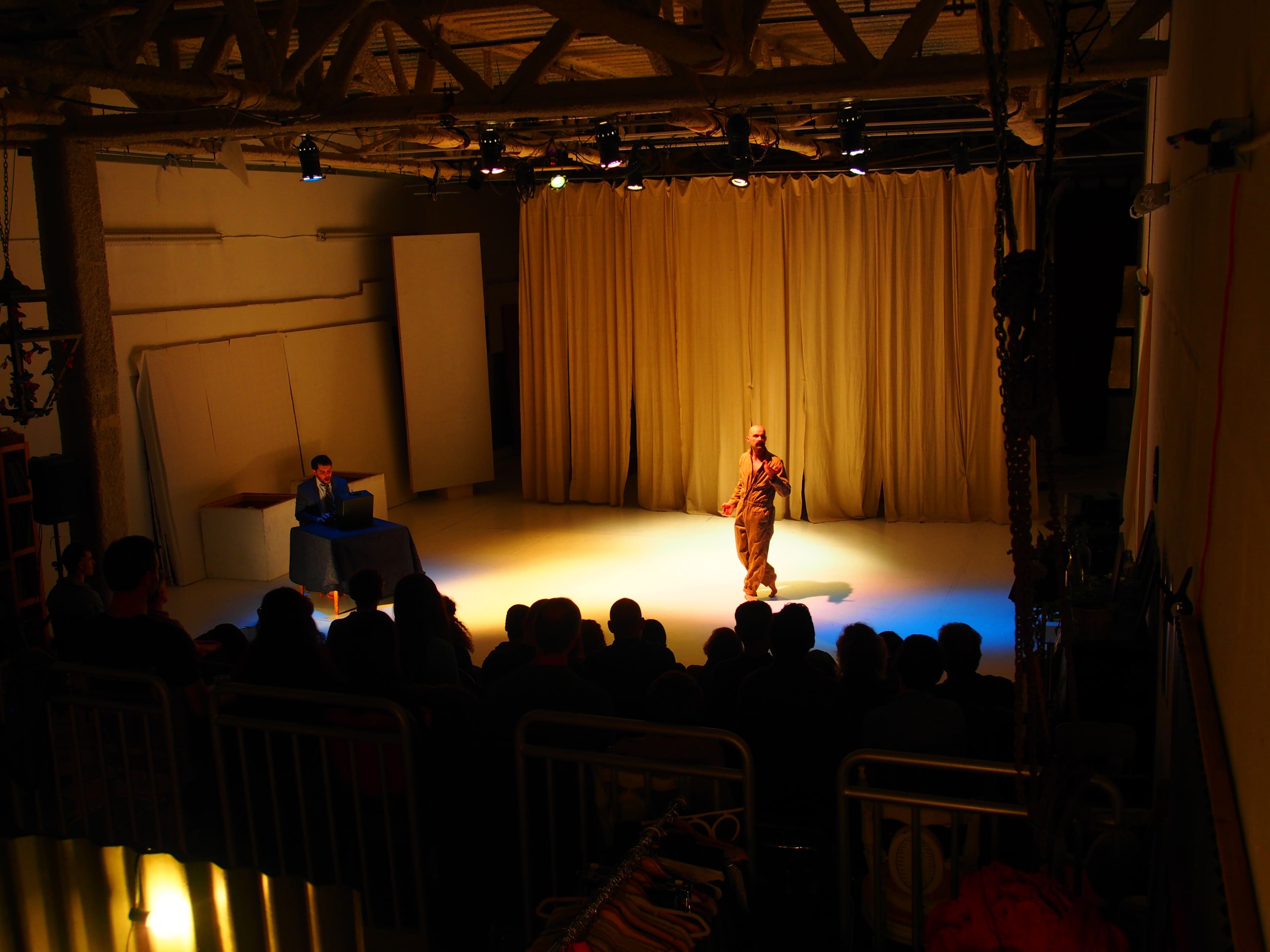 A performer stands on stage in front of a seated audience, while another individual is positioned at a table to the side, illuminated by stage lights.