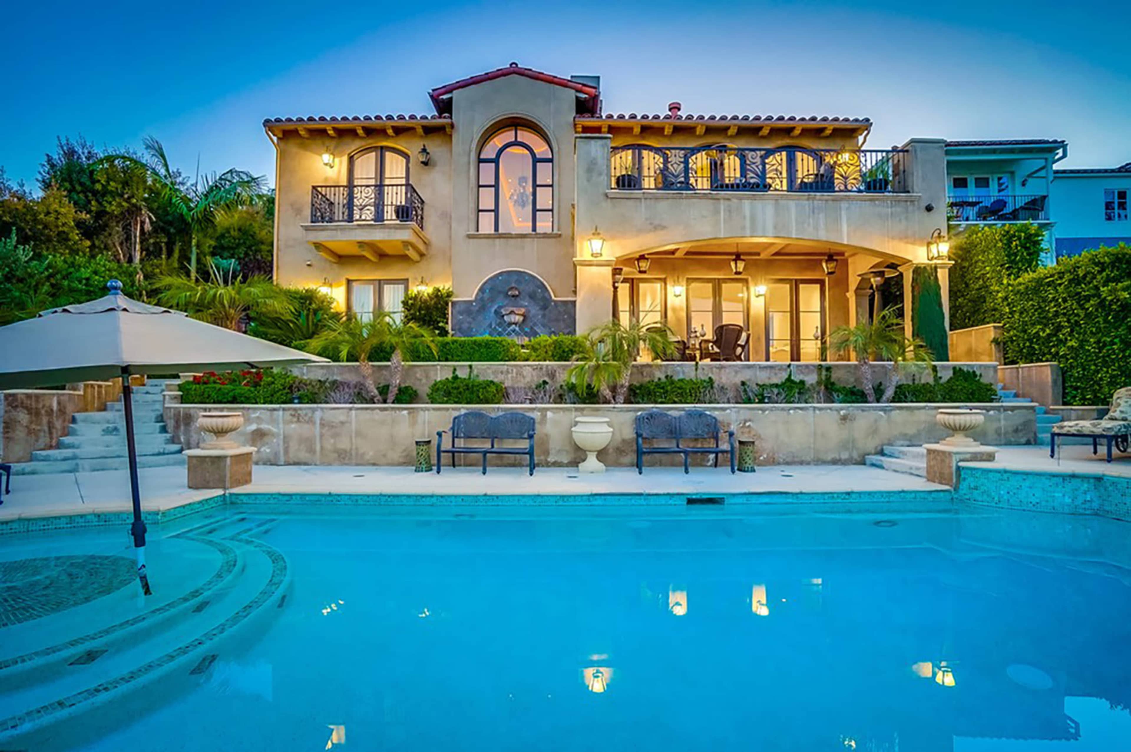 A two-story house with a balcony overlooking a swimming pool surrounded by landscaped gardens.