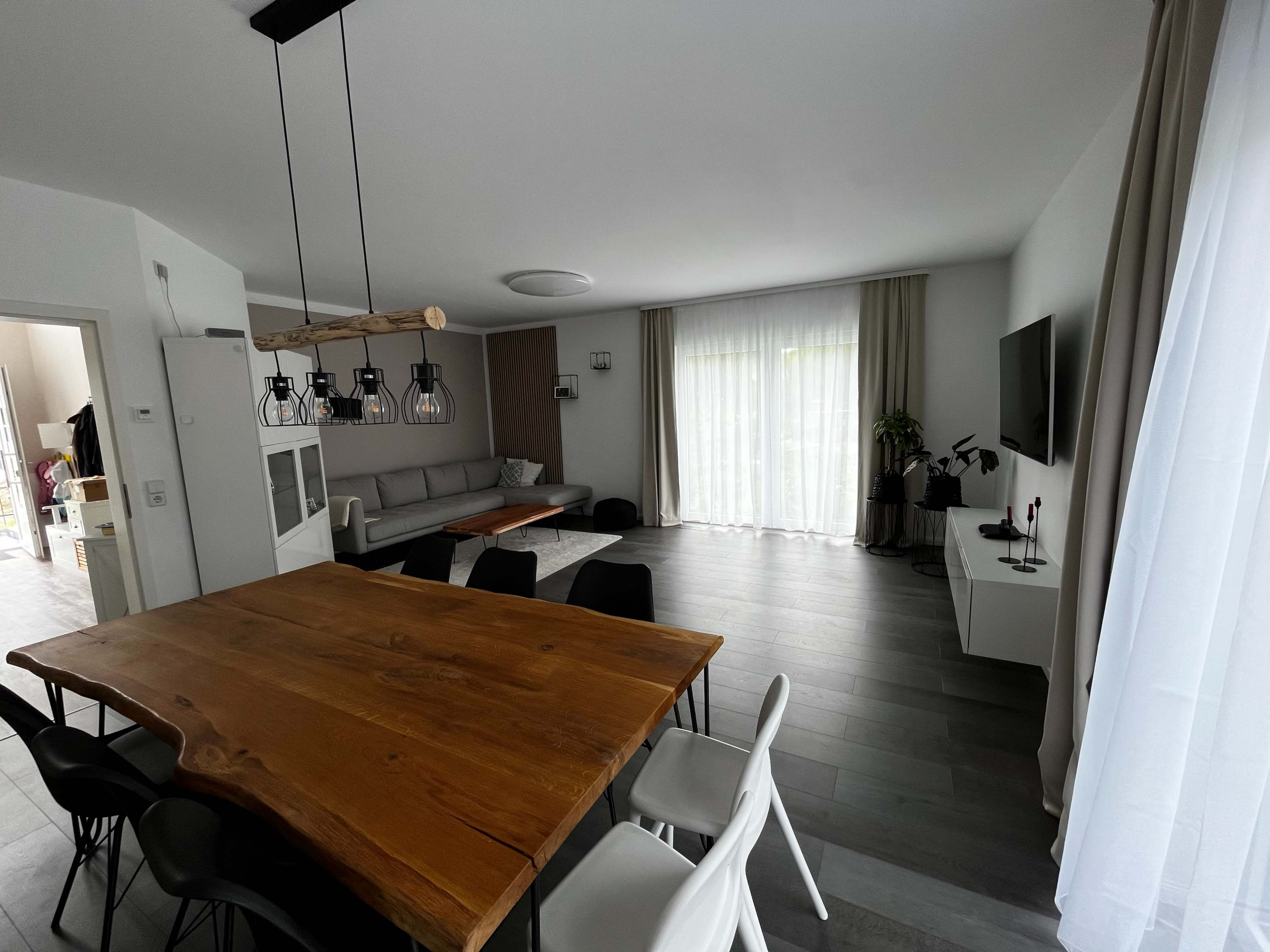 The image shows a modern living space featuring a wooden dining table, black and white chairs, a sofa, and large windows with sheer curtains.