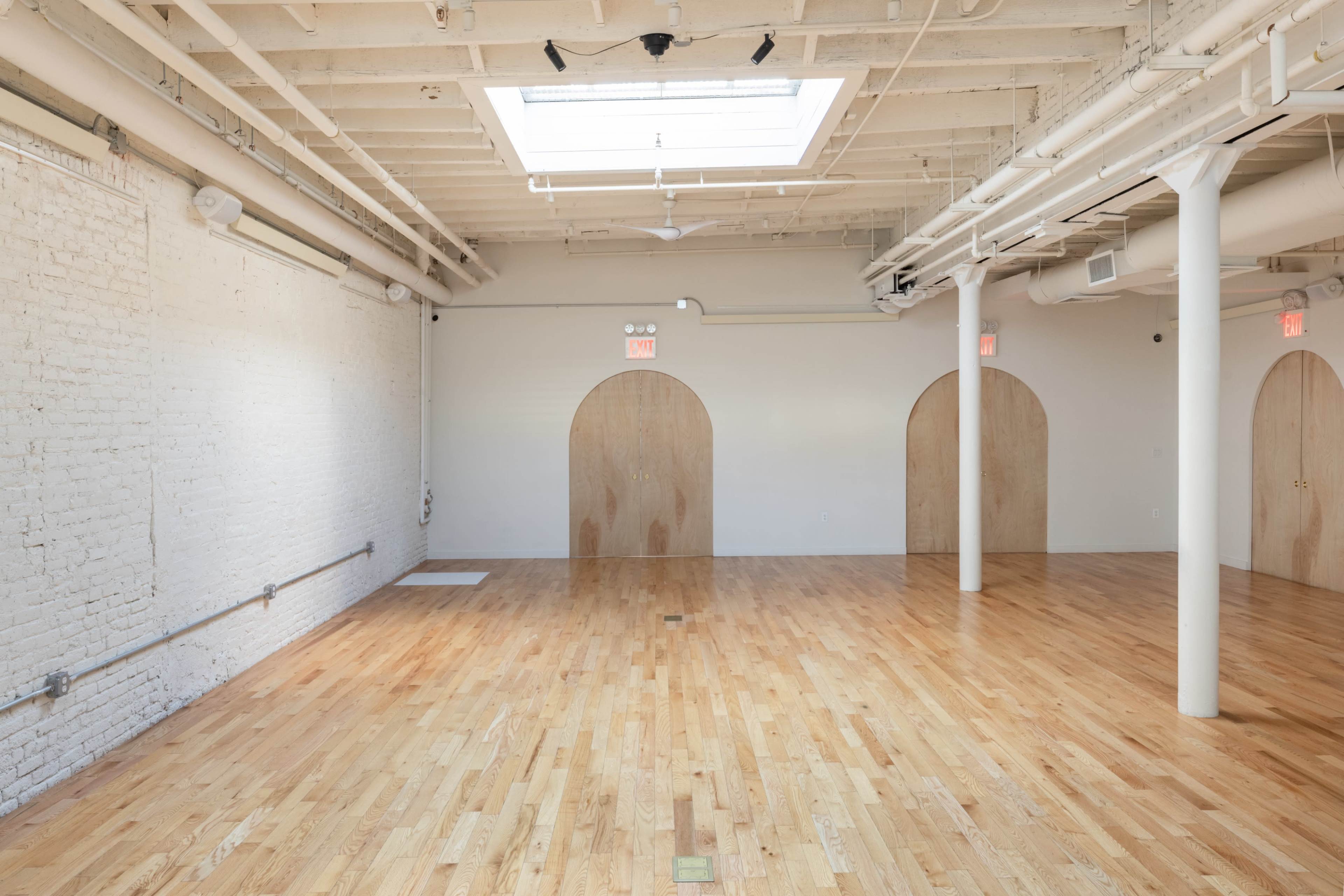 A spacious, empty room with polished wooden floors, exposed brick walls, and three archway entrances.