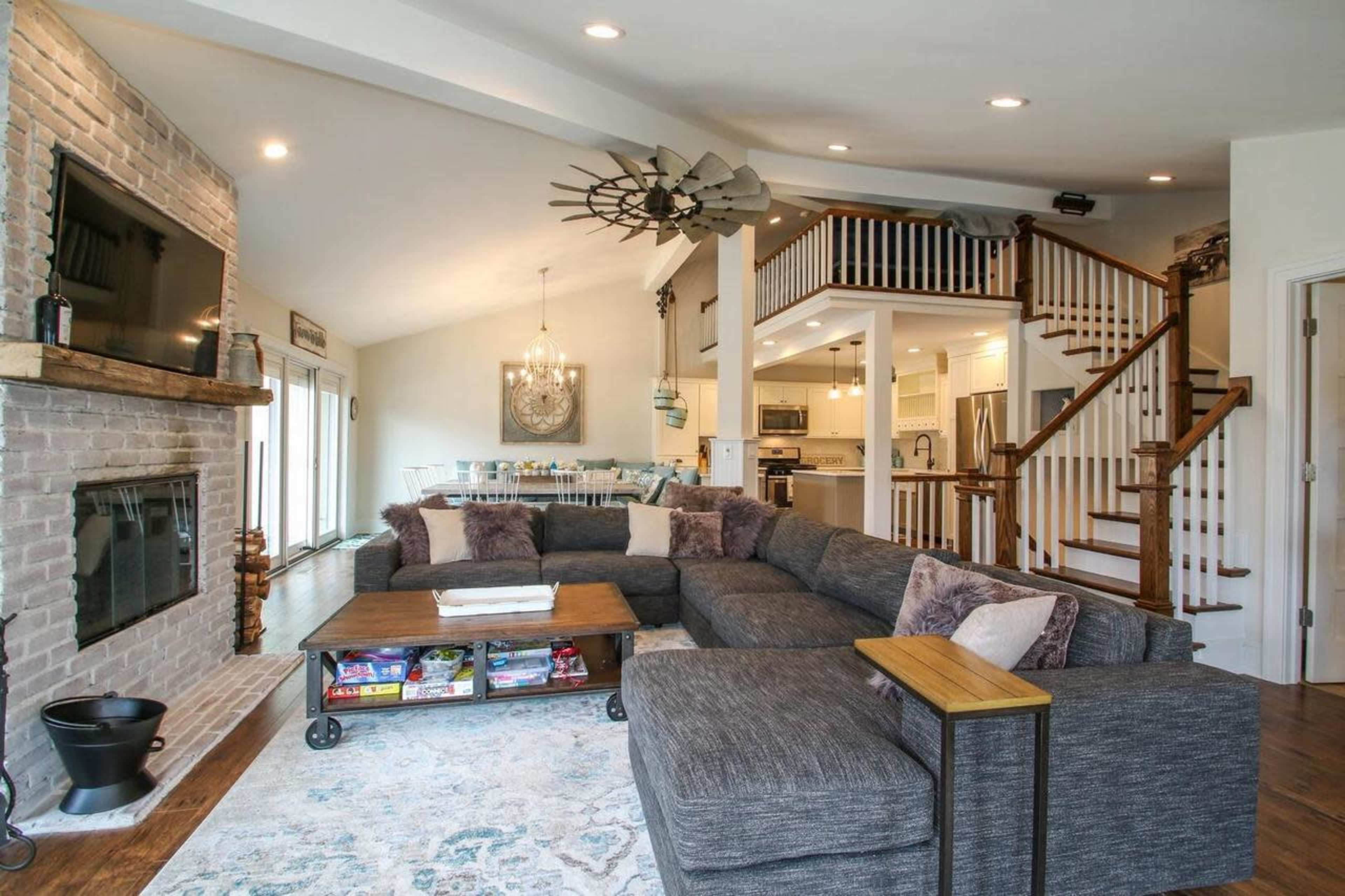 A spacious living room features a large sectional couch facing a brick fireplace and a flat-screen TV, with an open staircase leading to an upper level.
