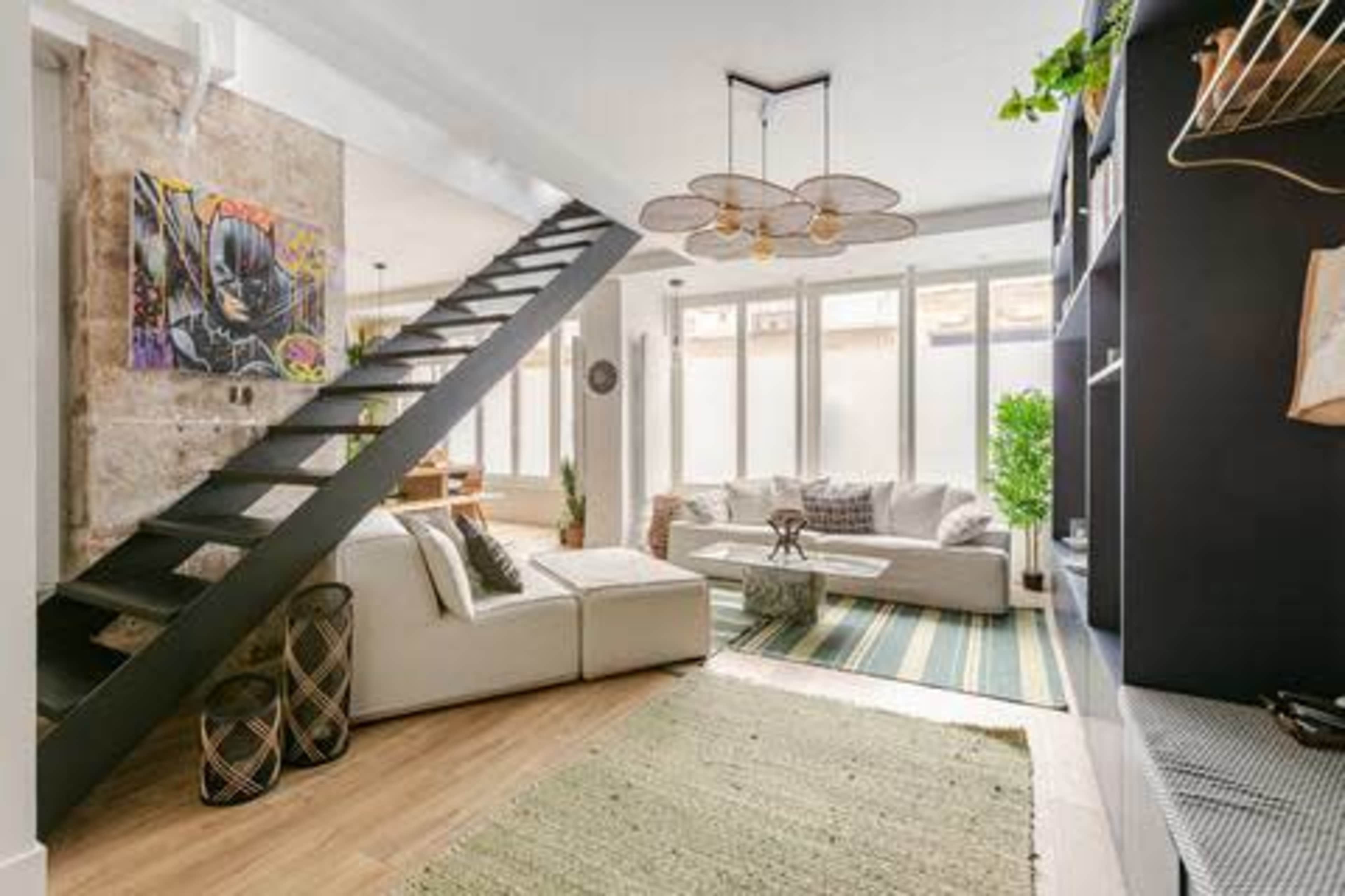 The image shows a modern living room with a sectional sofa, a staircase, and large windows allowing natural light to fill the space.