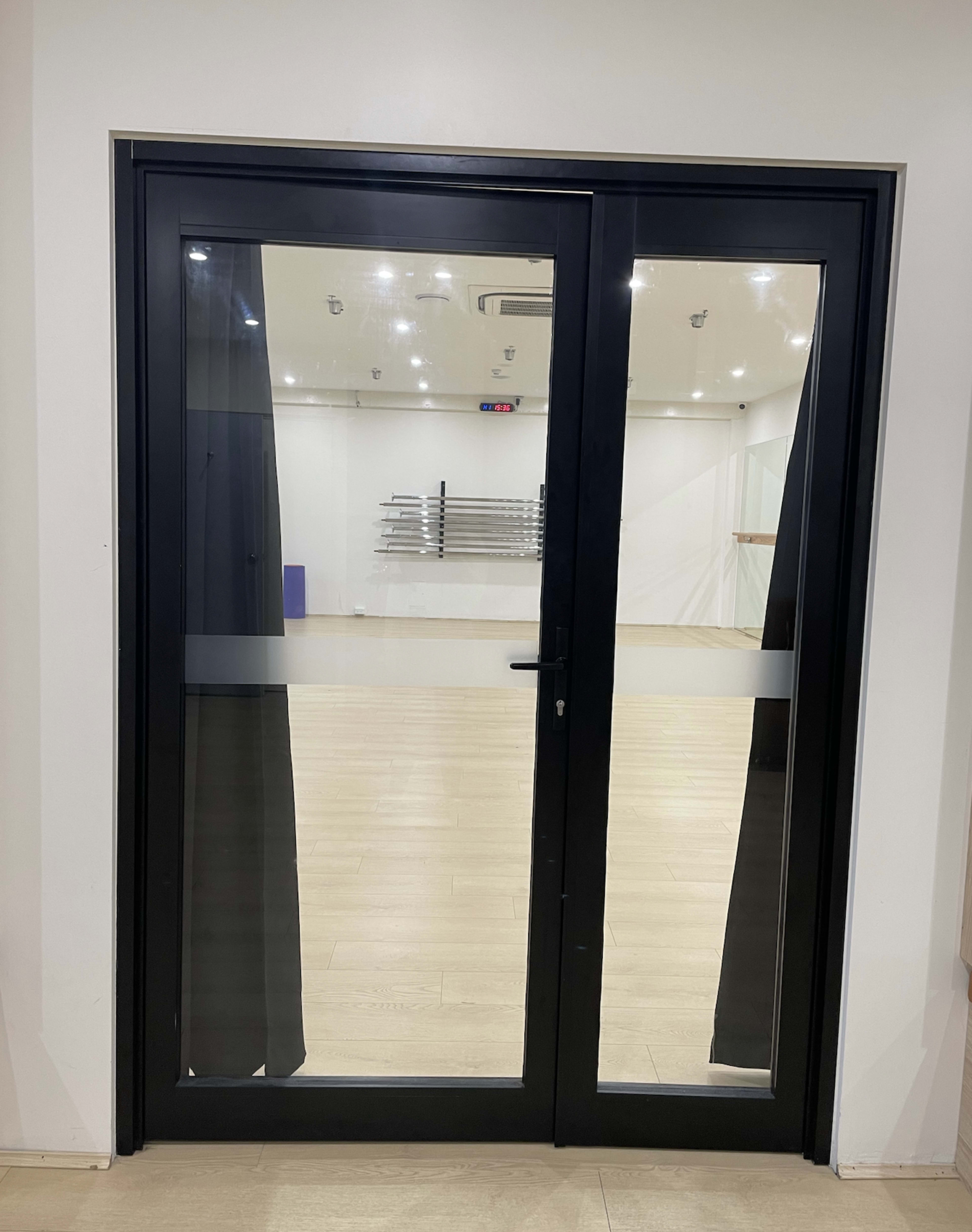 A black-framed double glass door leading into a brightly lit room with a wooden floor and equipment racks in the background.