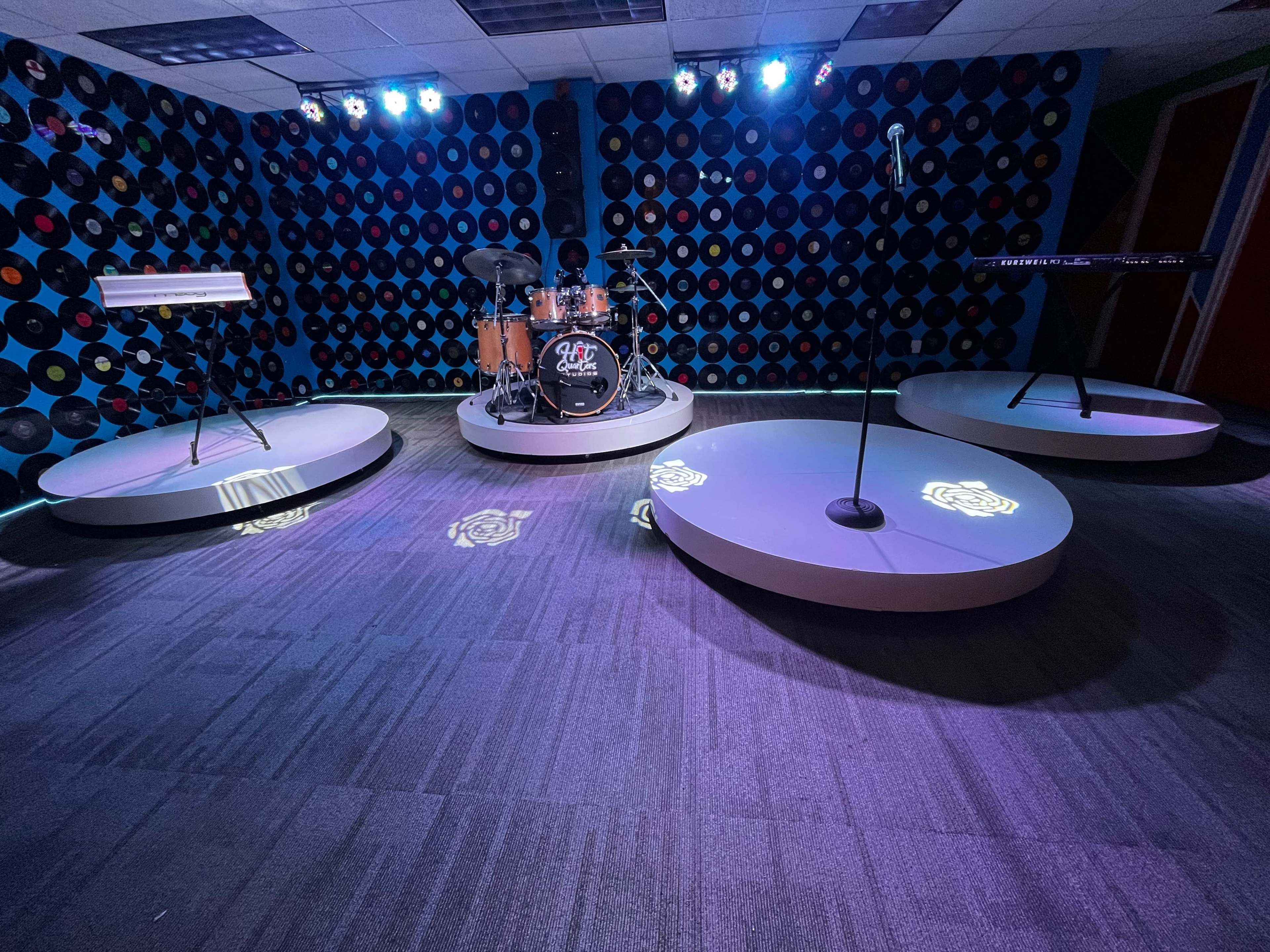 A music stage setup with a drum kit, two keyboards, and a microphone stand, all arranged on circular platforms illuminated by stage lights against a backdrop of vinyl records.