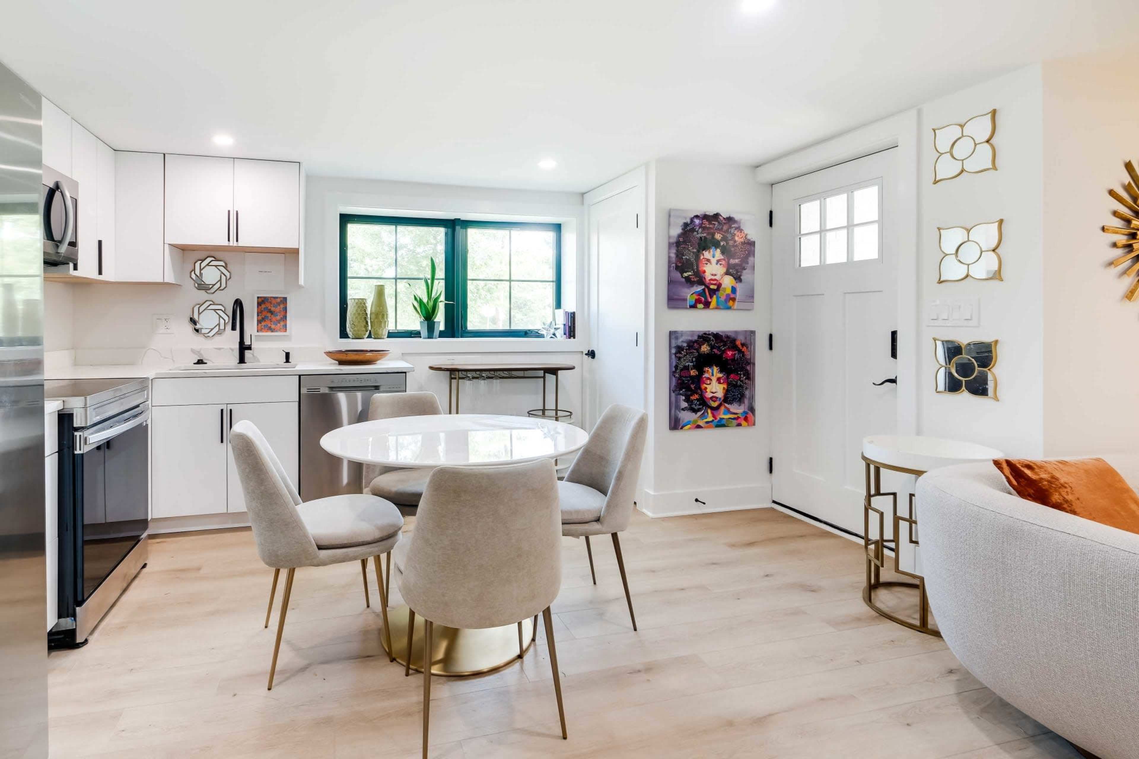A modern kitchen area with a circular dining table surrounded by gray upholstered chairs, next to a door and adorned with colorful artwork on the walls.