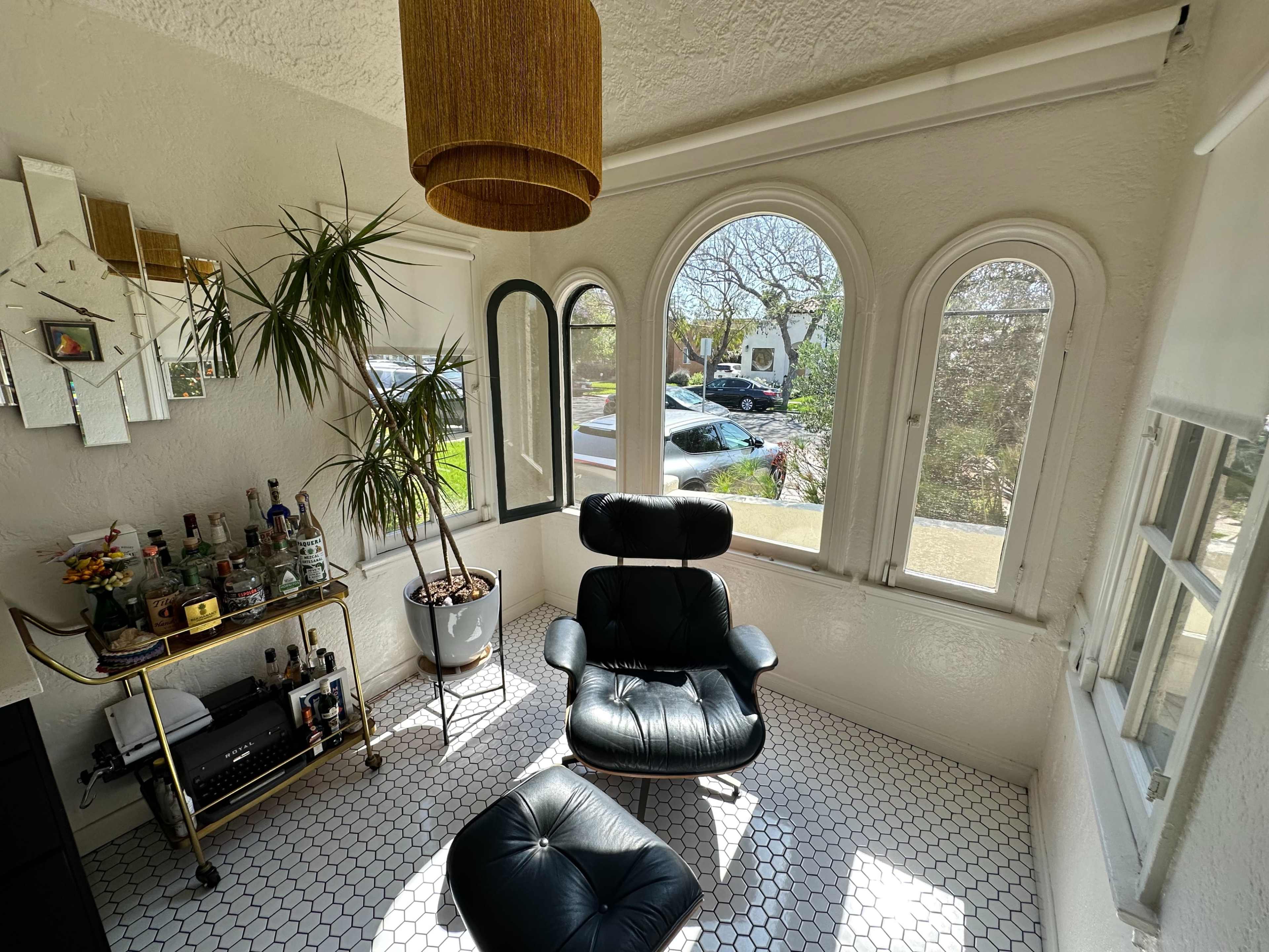 The image shows a sunlit corner of a room with a black leather chair, a bar cart, and large arched windows overlooking a garden and parked cars.