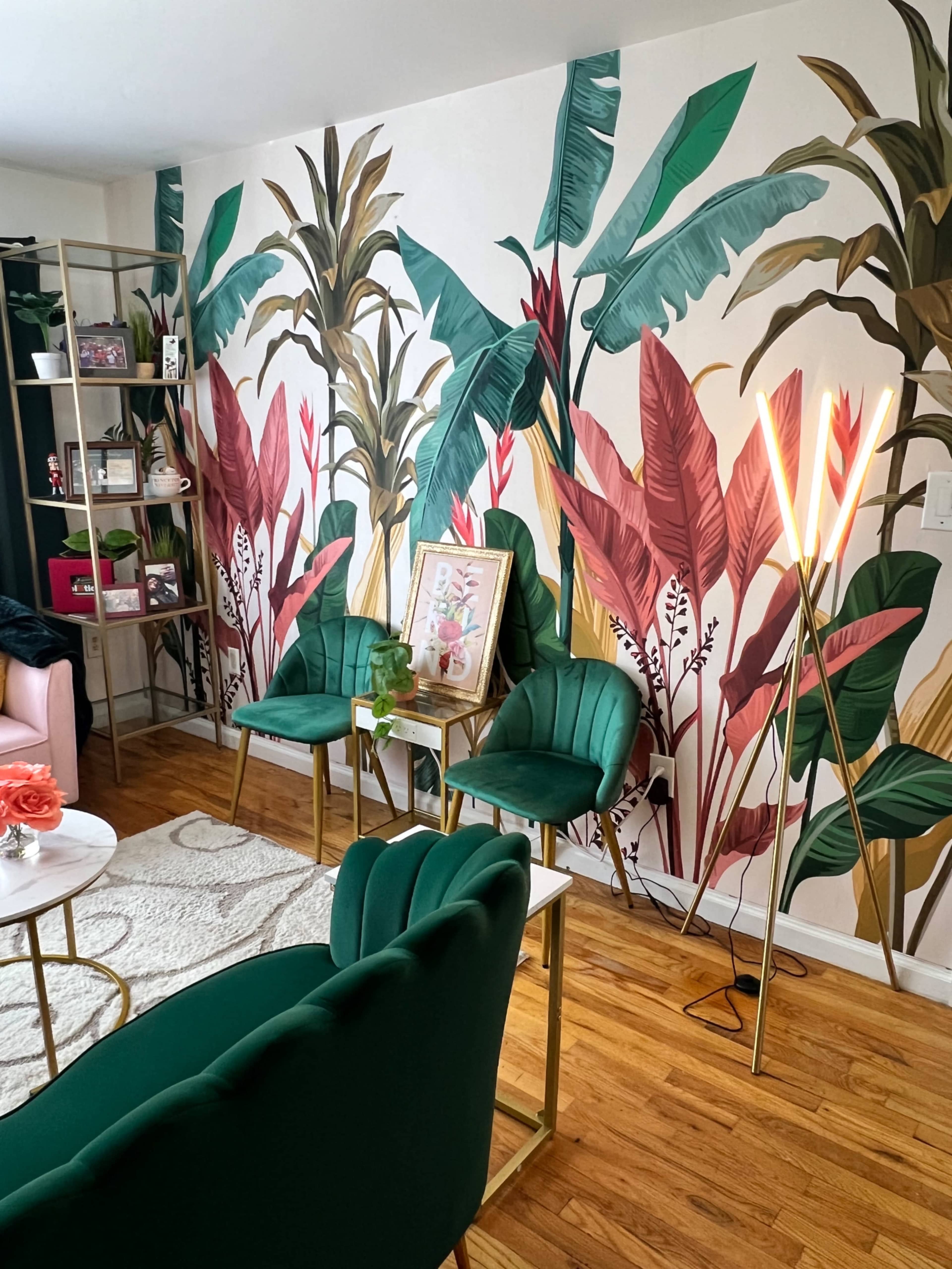 The image shows a living room with a tropical-themed mural on the wall, featuring green and red plants, complemented by green chairs and a gold floor lamp.