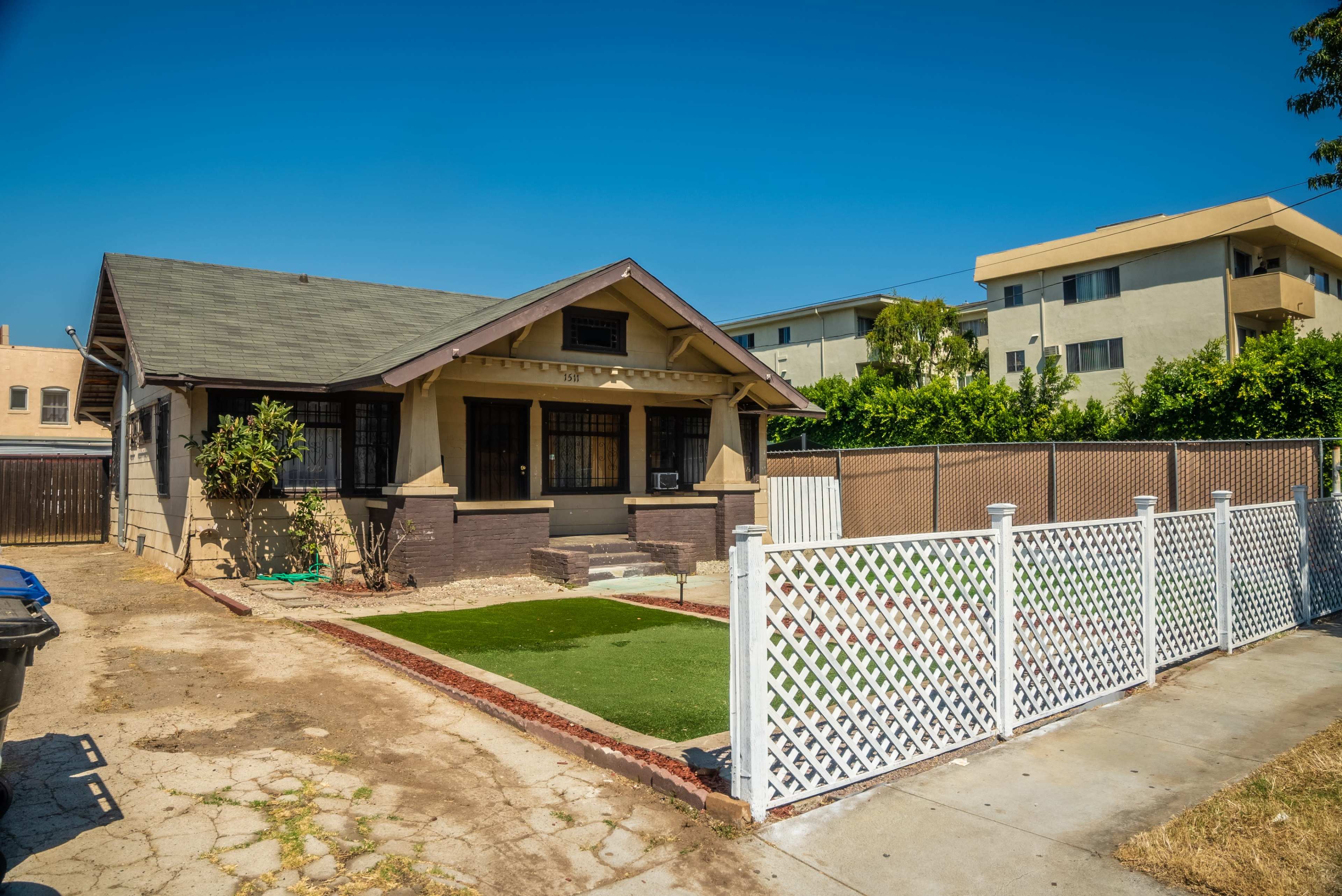 The image shows a single-story bungalow with a front yard enclosed by a white lattice fence, situated beside a multi-story apartment building.