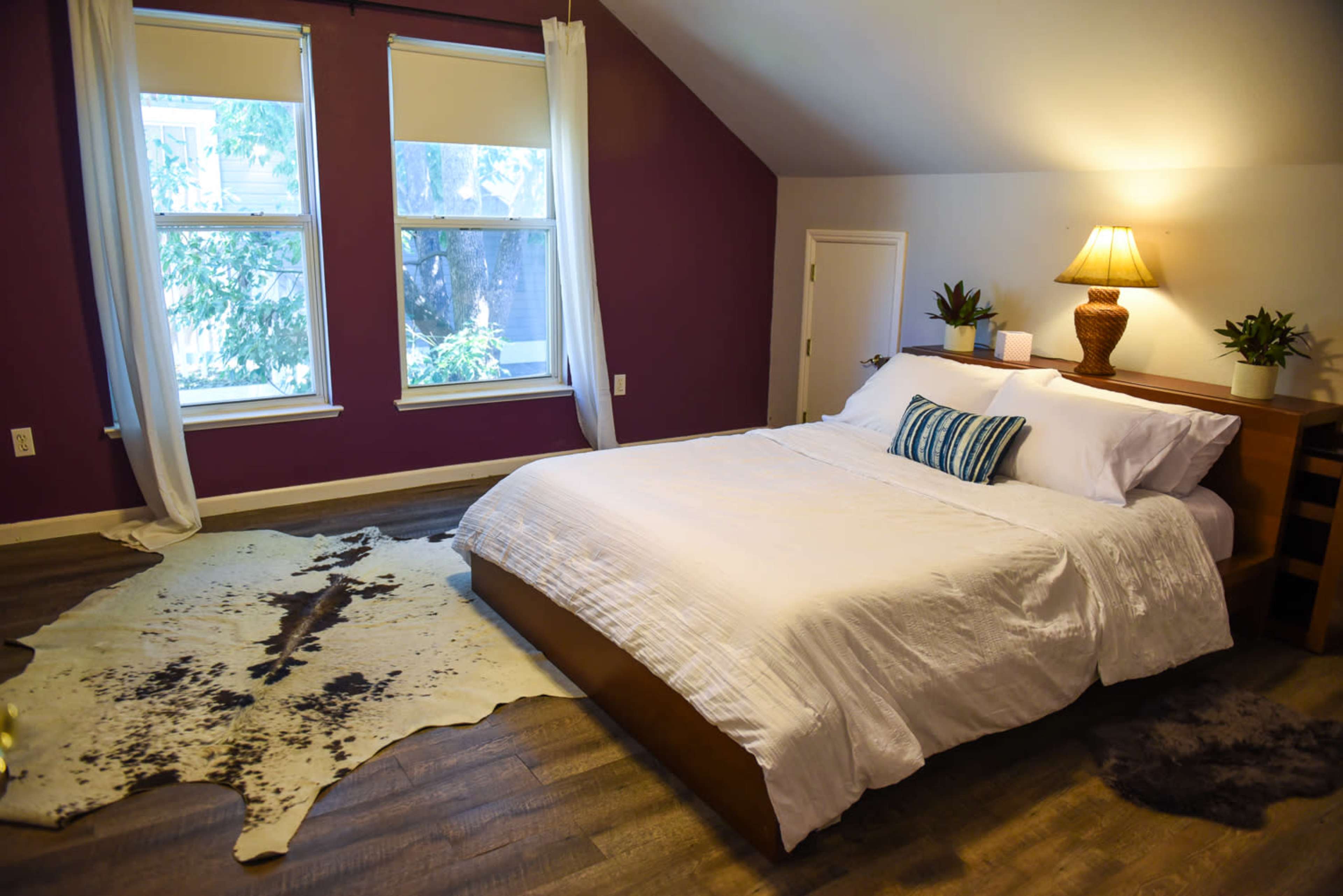A bedroom with a large bed, a nightstand with a lamp, and a cowhide rug on the wooden floor.