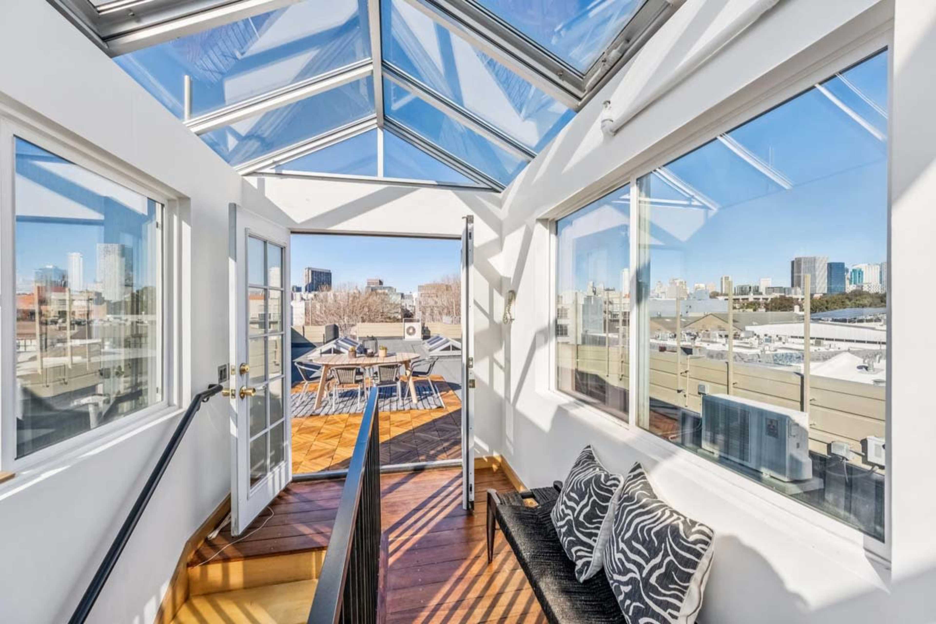 The image shows a bright indoor space with large glass windows and doors leading to a rooftop terrace, offering a view of a city skyline.