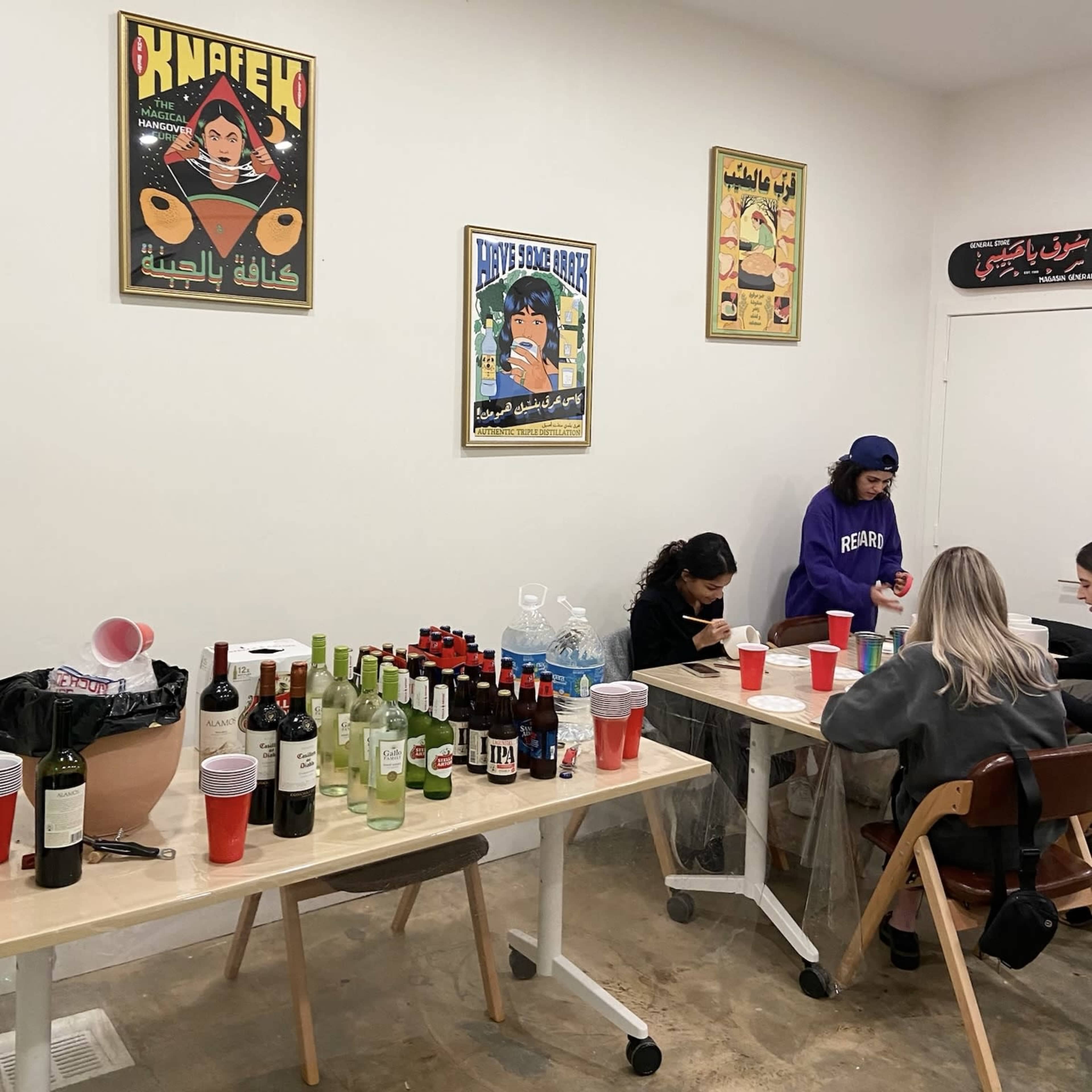 The scene shows a room with two tables covered in various bottles and drinks, while three people are seated, engaged in activities, and colorful posters decorate the walls.