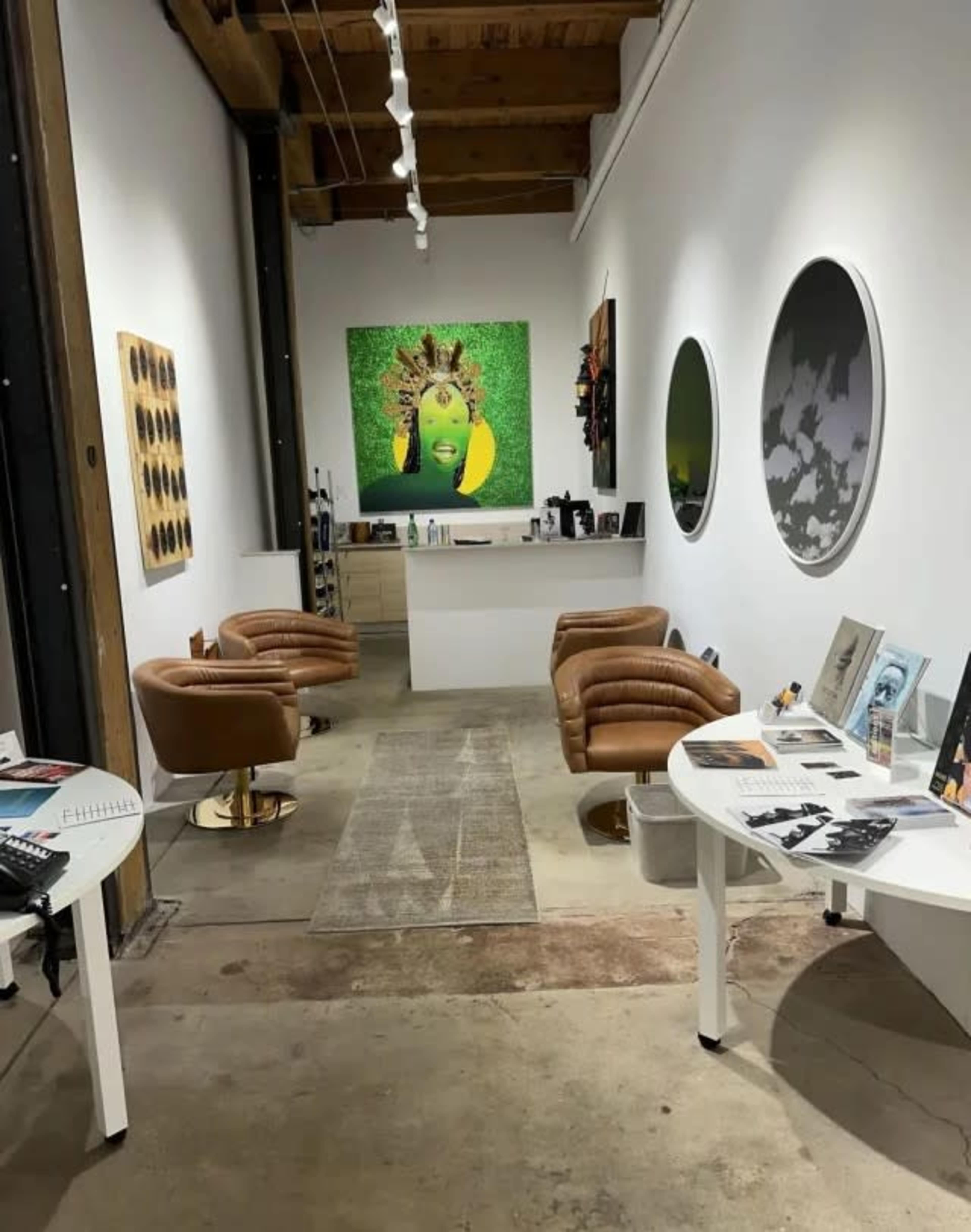The image shows a modern gallery space with two brown chairs, circular artworks on the walls, and a vibrant green piece featuring a face at the far end.