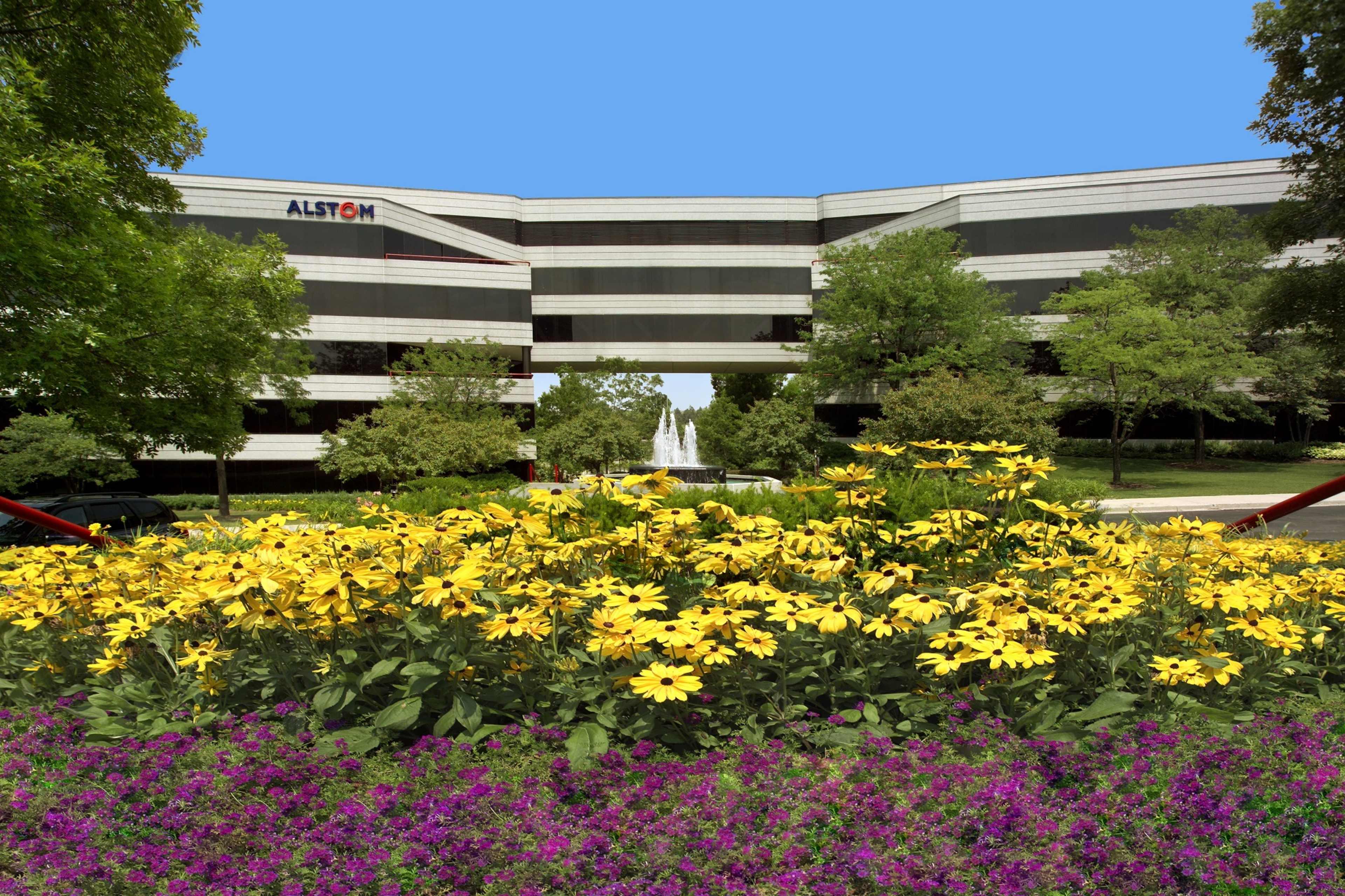 A modern office building with the name "ALSTOM" is framed by vibrant flower beds in the foreground and surrounded by greenery.